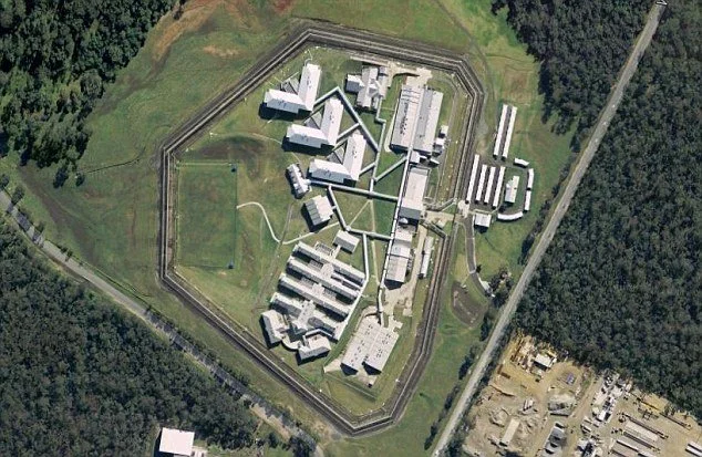 Aerial view of a high-security prison surrounded by a tall fence with guard towers, multiple buildings inside the compound, and a few roads outside the perimeter.