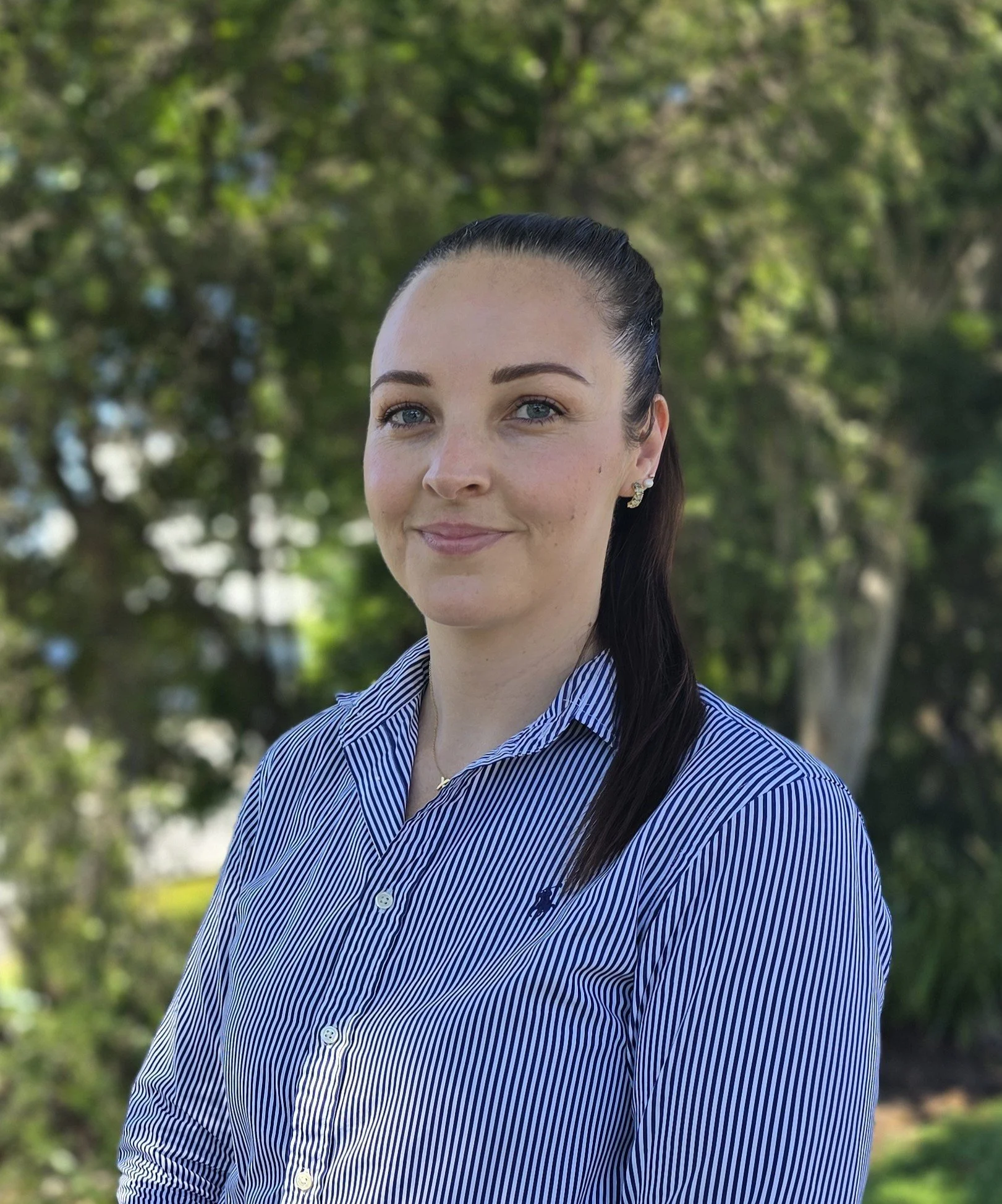 A woman with dark hair pulled back, wearing a blue and white striped shirt, posed outdoors with trees in the background.
