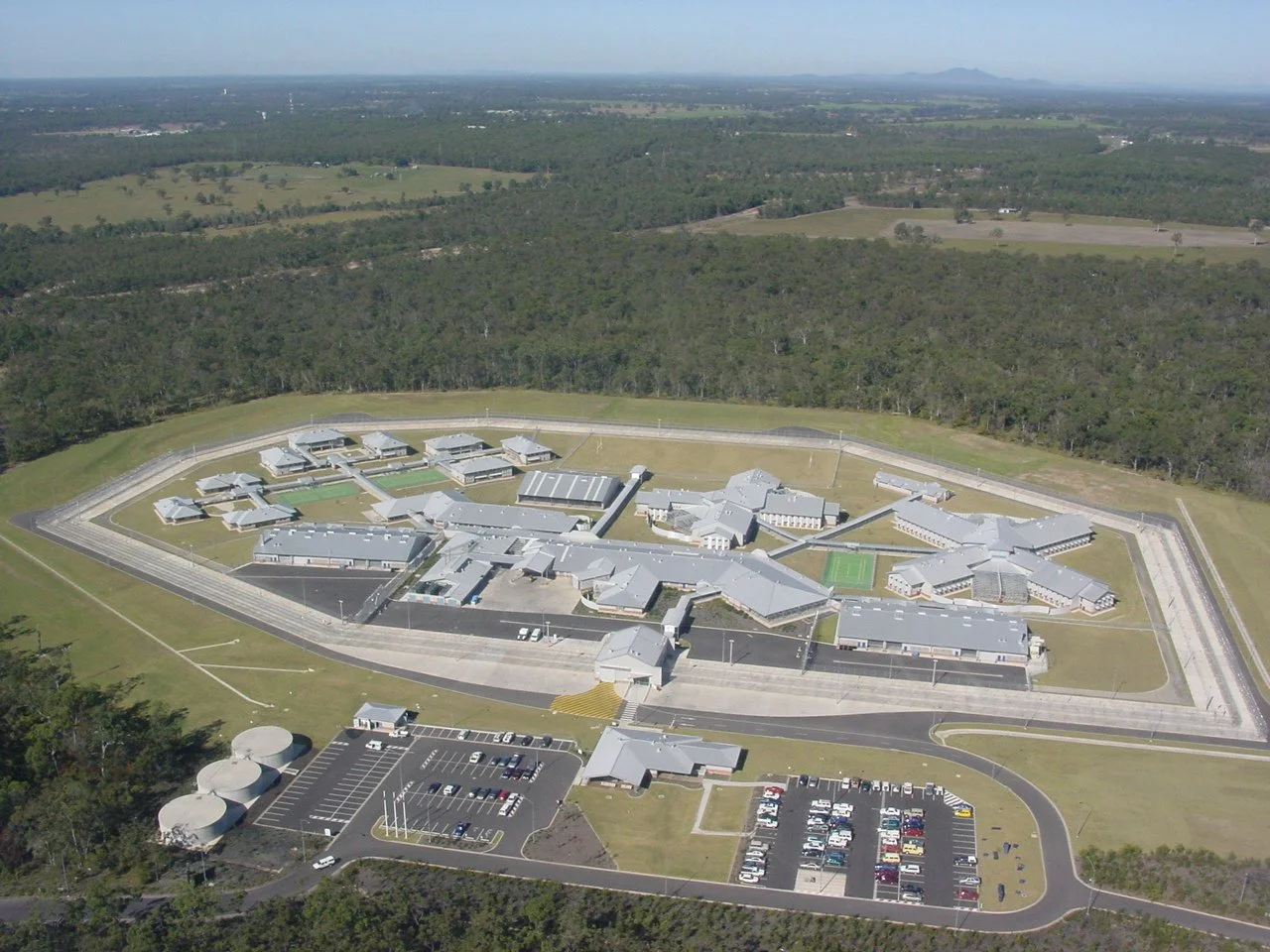 Aerial view of a large prison or correctional facility with multiple buildings, surrounded by a security fence and a parking lot, situated in a rural area with fields and forested land in the background.