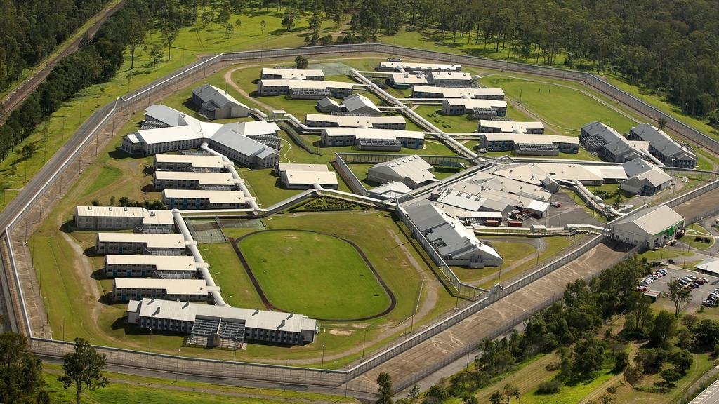 A large correctional facility, with multiple interconnected buildings surrounded by high fences and monitored by security cameras, set in a green, wooded area.