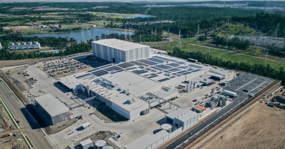 Aerial view of a large industrial facility with white buildings, parking lot, and surrounding green landscape.