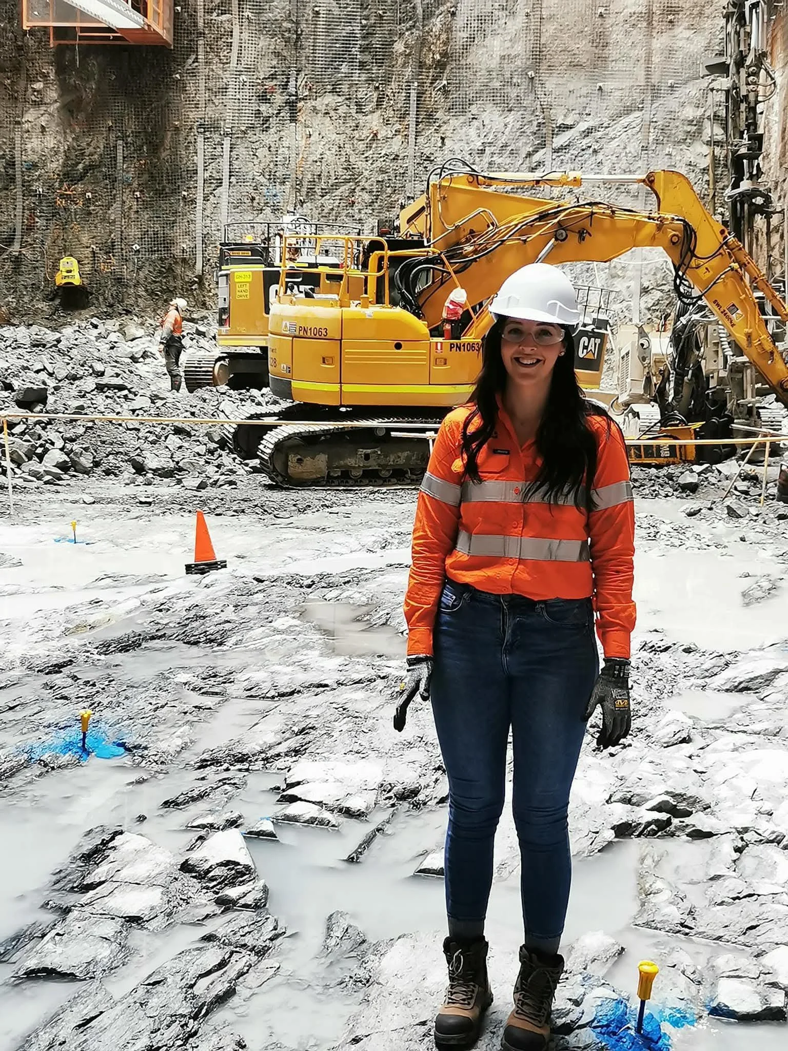 A woman wearing safety gear, including a white helmet, orange high-visibility shirt, jeans, and work boots, standing inside a construction site with a large yellow excavator and rocky terrain in the background.