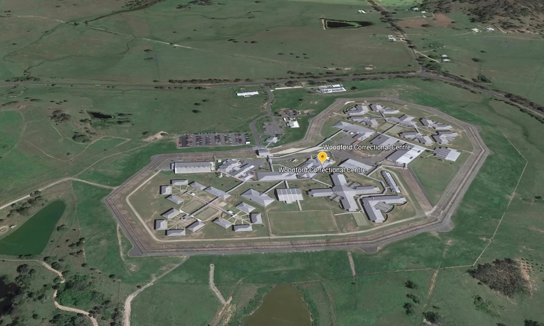 Aerial view of Woodford Correctional Centre, a large prison complex surrounded by green fields, roads, and ponds.