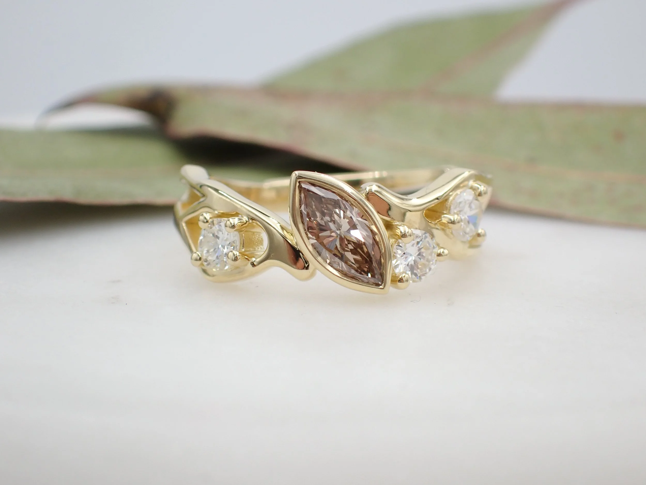 Yellow gold Kintsugi inspired ring set with an Argyle marquise central diamond in a bezel setting accompanied with smaller white round diamonds.  Ring is laying on a flat white surface with gum leaves in the background.