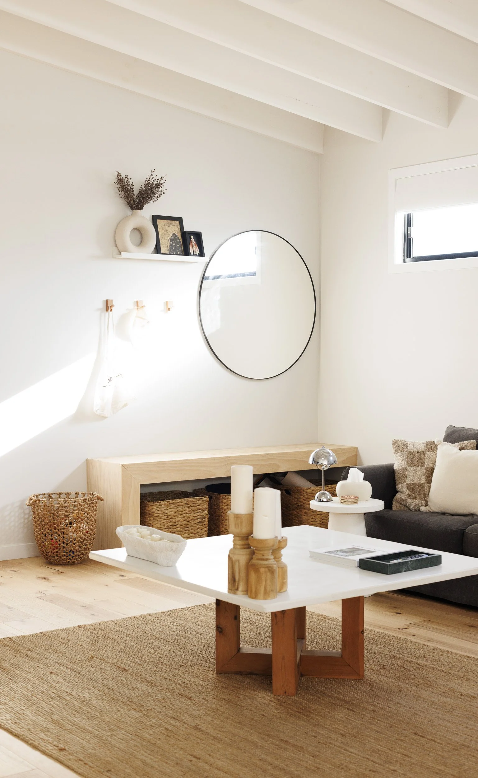 Minimalist living room with white walls, wooden floor, a white coffee table with candles, a black couch with cream and checkered pillows, a woven basket, a wall shelf with decorative items, a round mirror, and a small window letting in natural light.