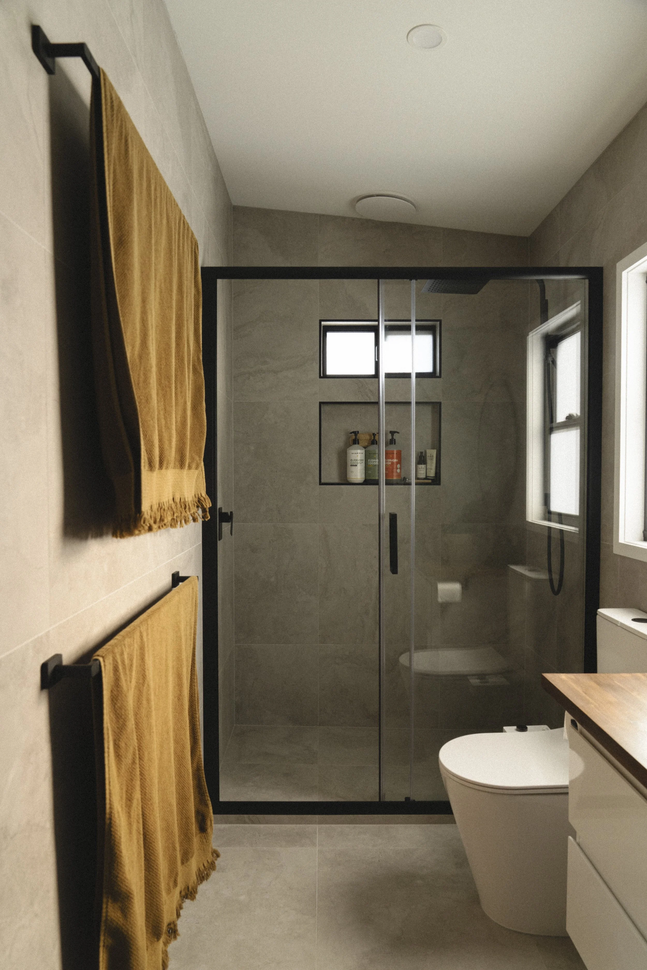Modern bathroom with a glass shower, beige tiles, two small high windows in the shower, yellow towels on black racks, a white toilet, and white cabinetry with a wooden countertop.