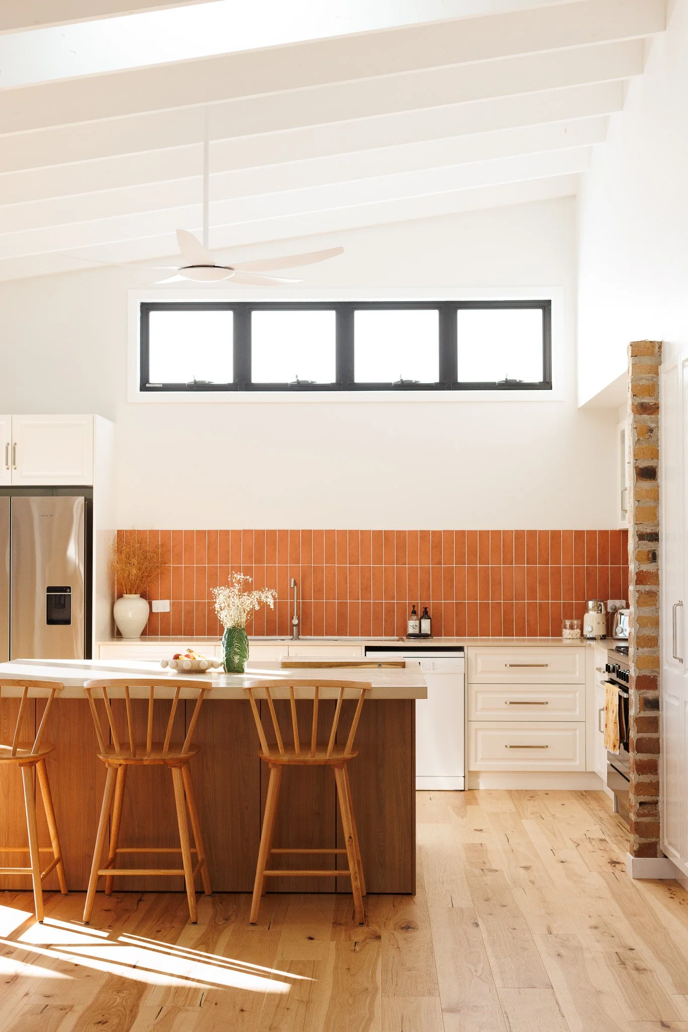 Modern kitchen with white cabinets, wooden island and bar stools, a reddish-brown tile backsplash, and a row of high windows near the ceiling, with hardwood flooring and a brick column on the right.