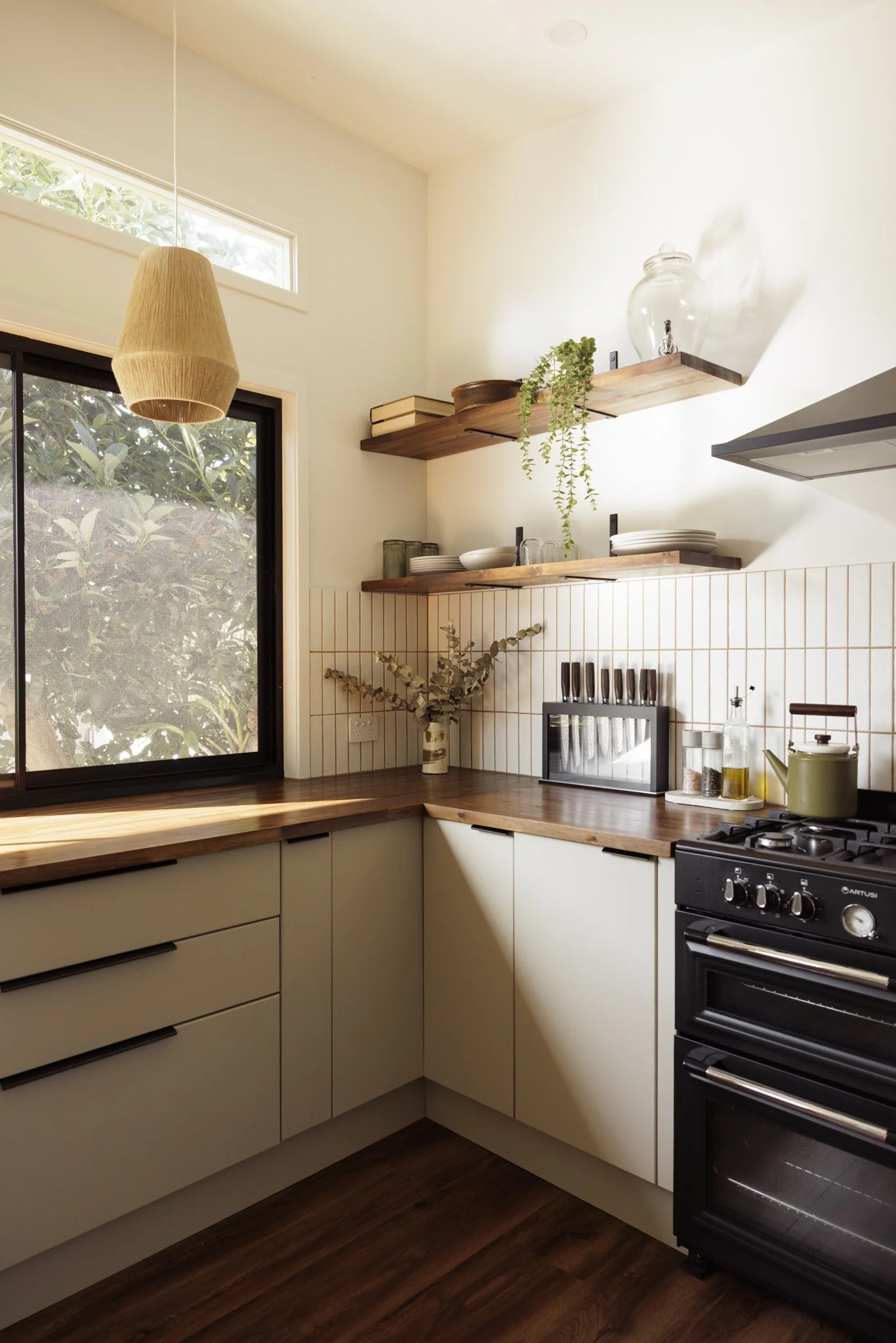 Kitchen with white cabinets, wooden countertops, open wooden shelves holding plates, bowls, books, and a glass jar, a window with greenery outside, and a black stove with a green kettle on top.
