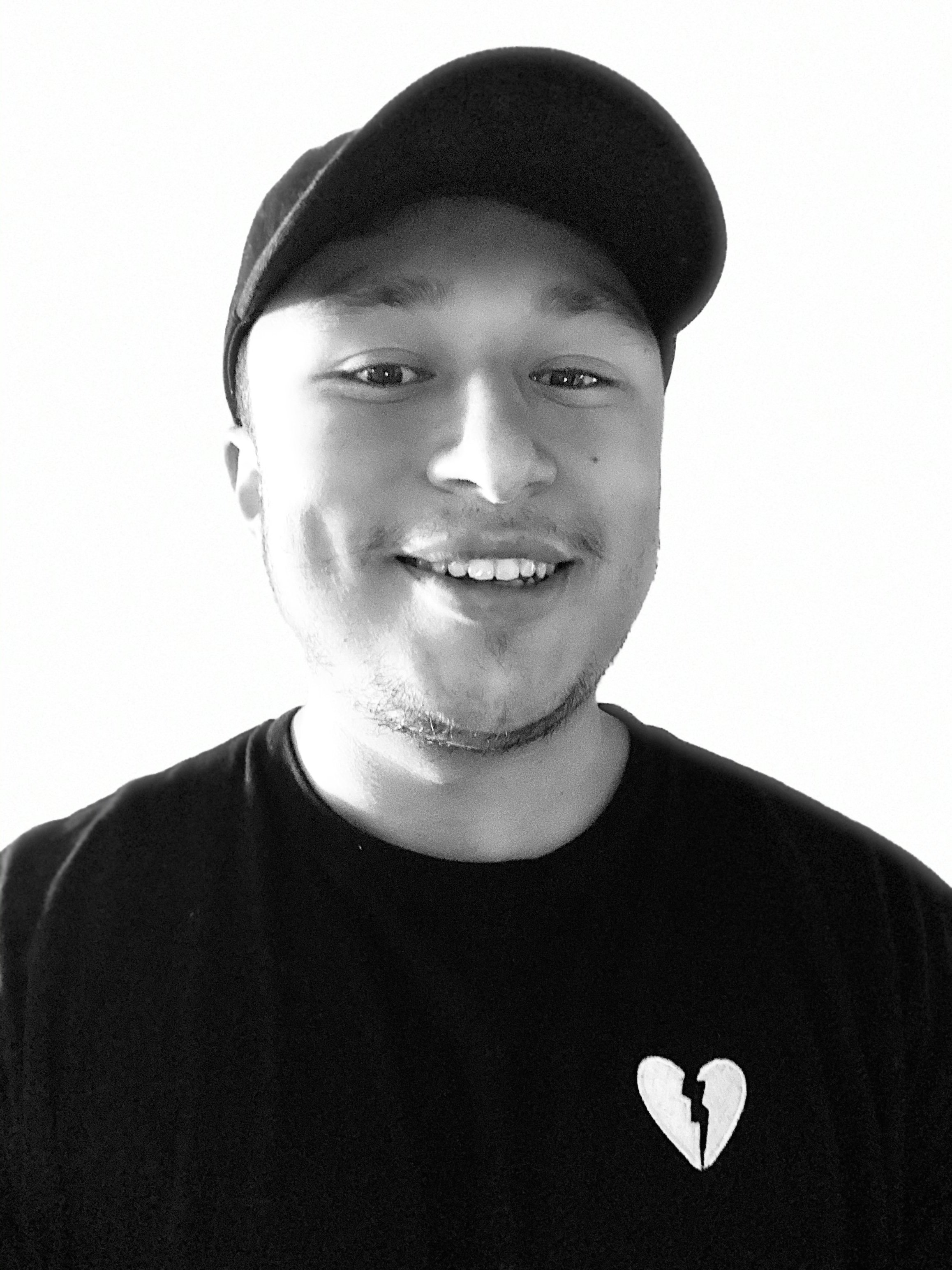 Black-and-white photo of a young man wearing a baseball cap and a T-shirt with a broken heart design, smiling at the camera.