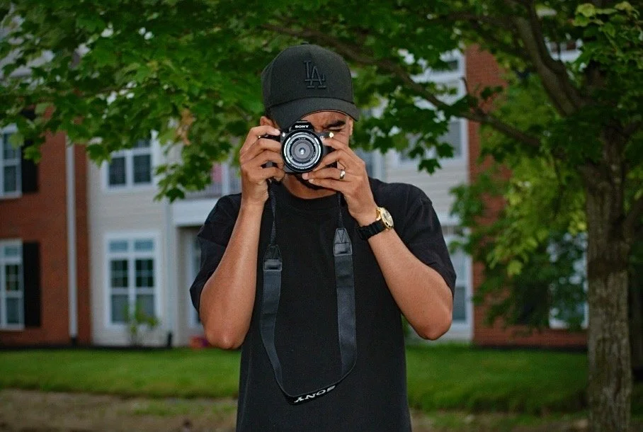 A man wearing a black cap and shirt, holding a camera up to his face and taking a photo outdoors, with trees and buildings in the background.