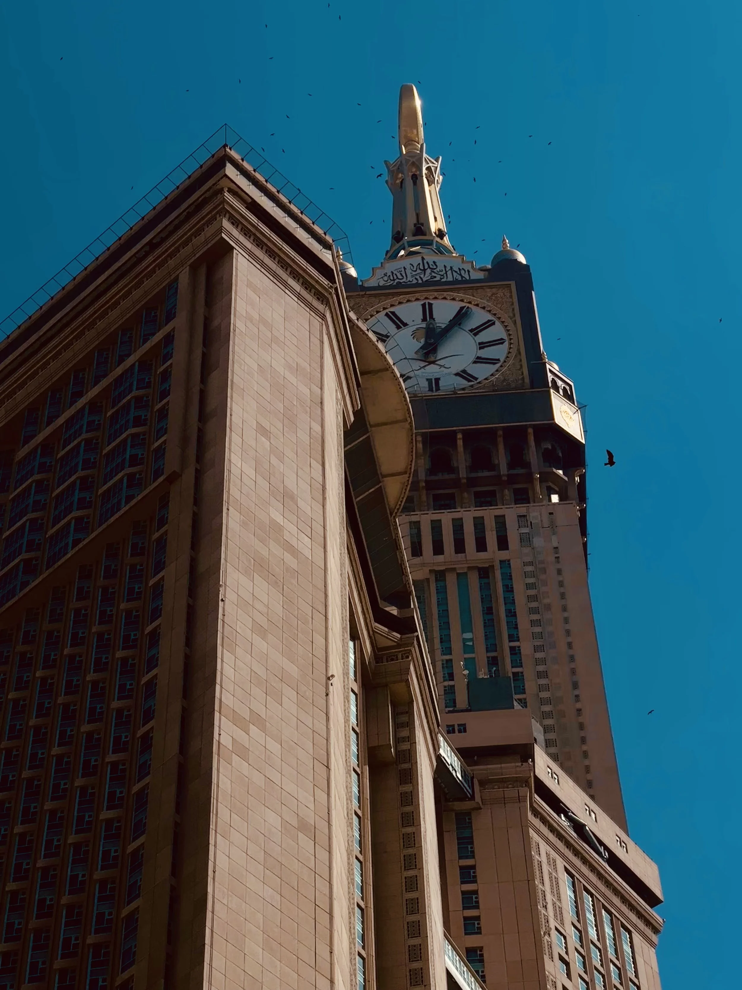 A tall clock tower with a golden spire and Arabic script near the clock face, against a clear blue sky with flying birds.