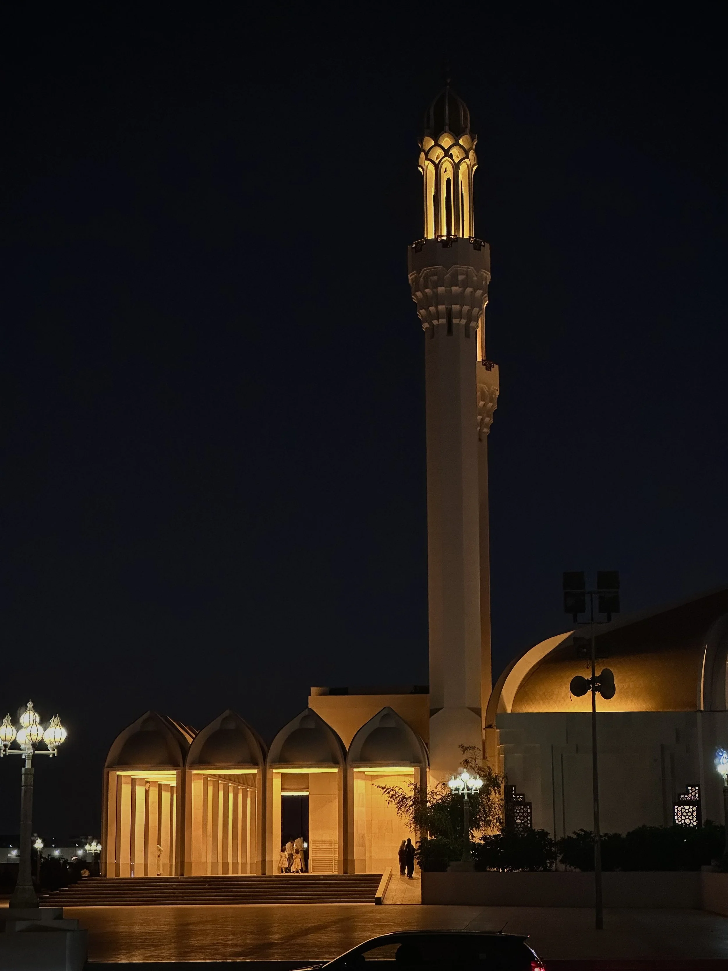 Night view of a mosque with illuminated domed arches and a tall minaret, with some people at the entrance and street lamps nearby.