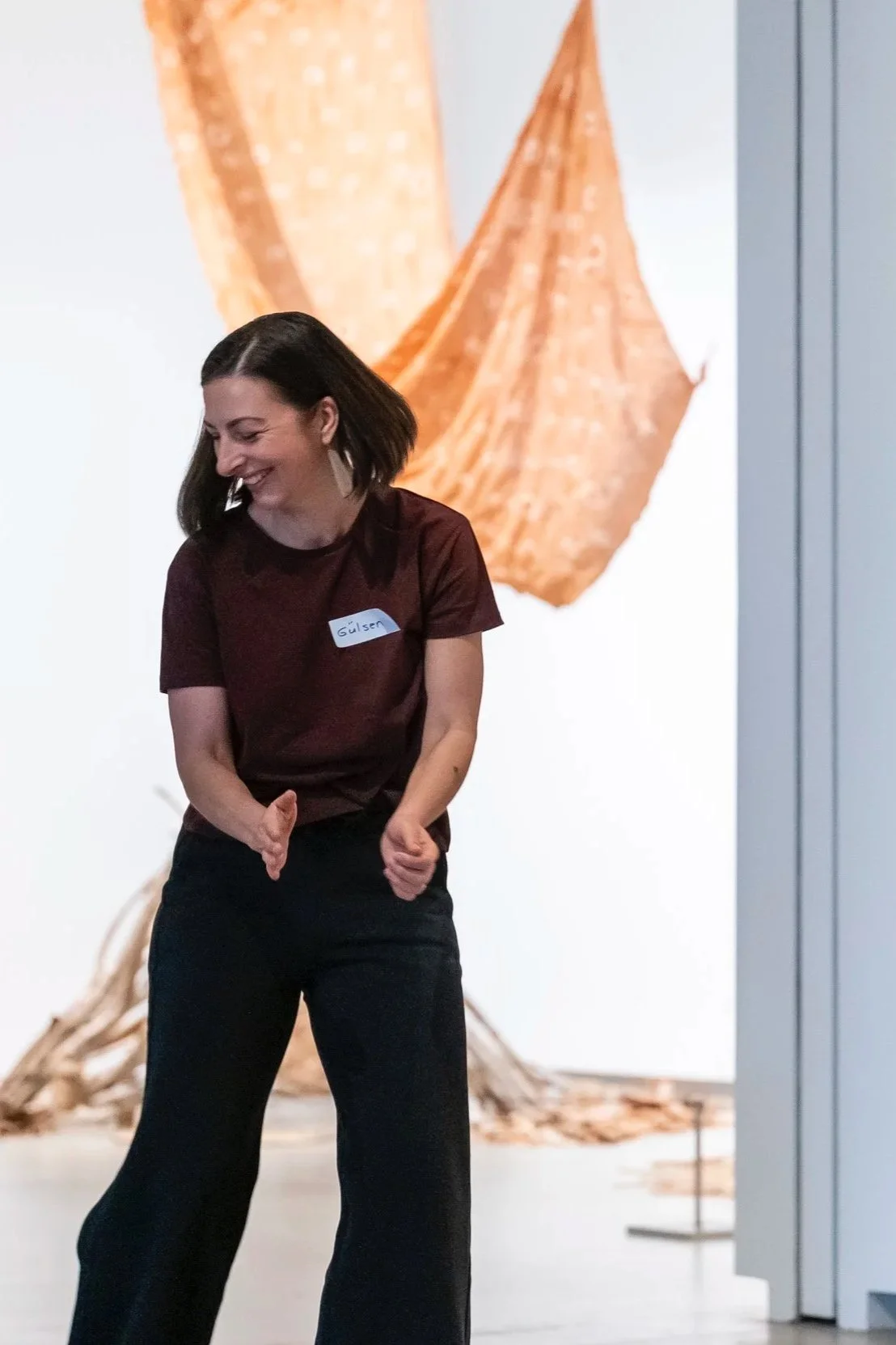 A woman with dark hair and wearing a maroon shirt and black pants, smiling and giving a thumbs-up gesture, standing in front of a large abstract artwork with orange and beige tones.