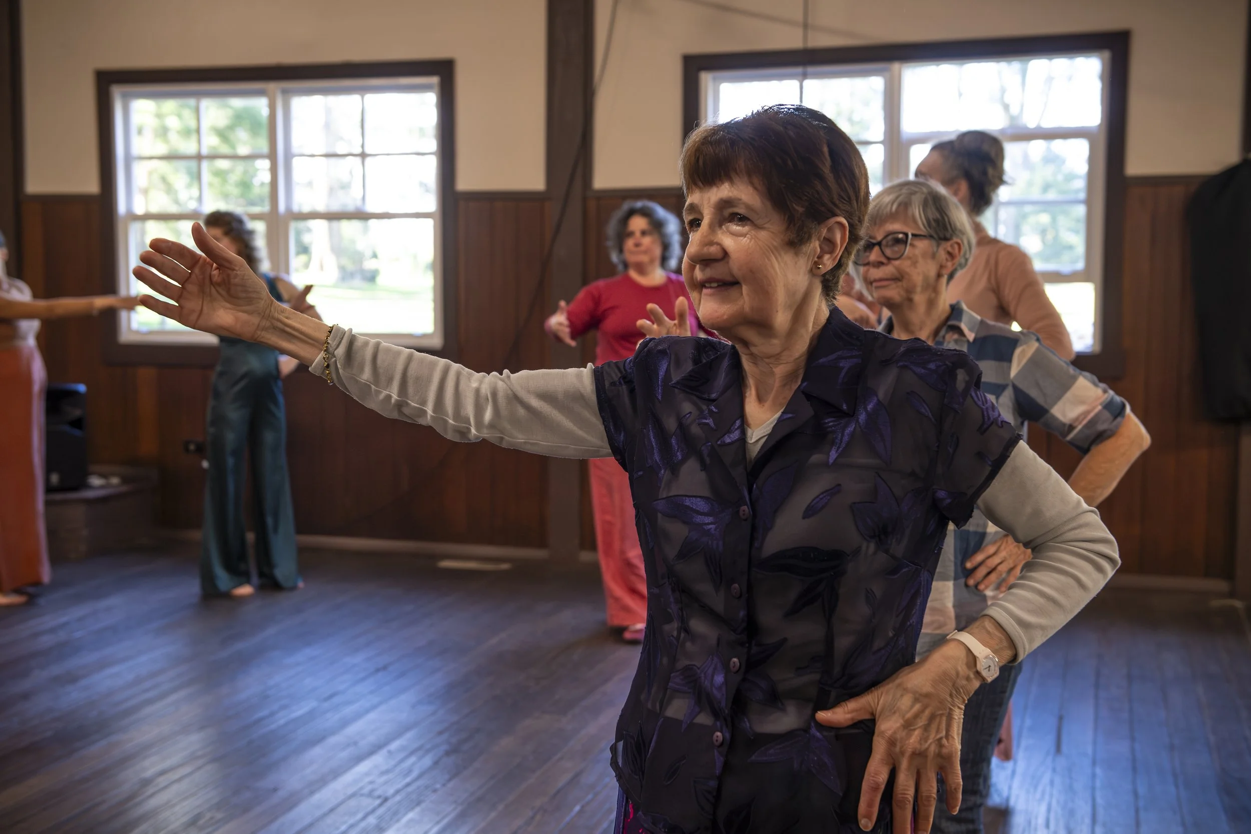Older women participate in a dance or exercise class indoors, with large windows showing green trees outside. One woman in the foreground extends her arm and smiles, surrounded by other women in casual clothing, engaging in stretching or movement activities.