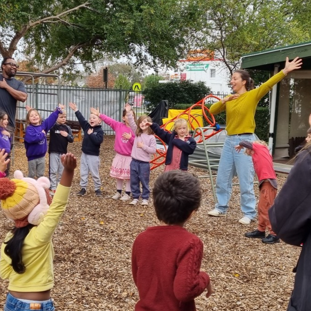 Children participating in an outdoor activity with an adult instructor, standing on a wood chip playground, with trees and playground equipment in the background.