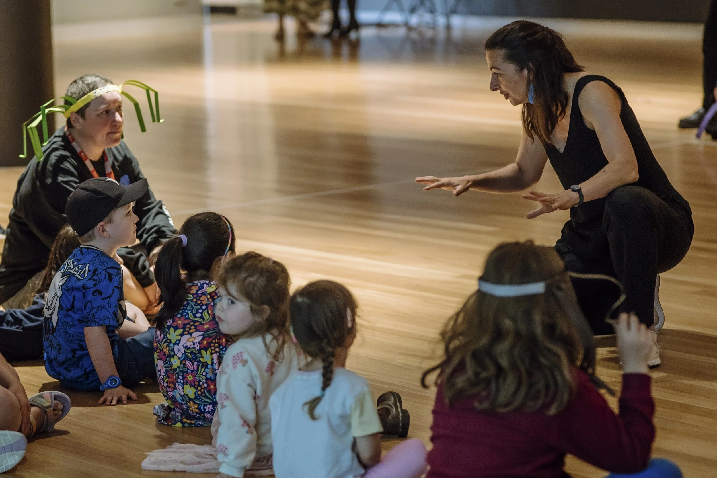 A woman kneeling and speaking to a group of children sitting on the floor. The children are listening attentively. The setting appears to be indoors with wooden flooring.