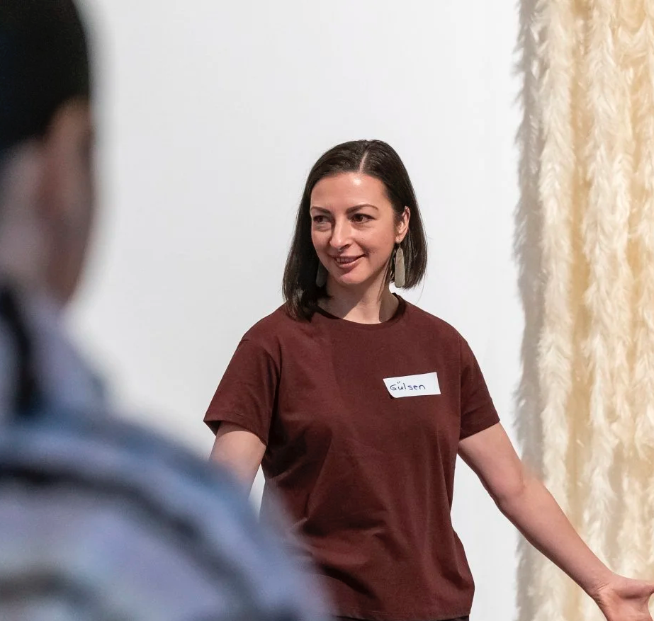 A woman with dark hair and earrings, wearing a maroon t-shirt, smiling and interacting with people in a room with a white wall and beige curtain.