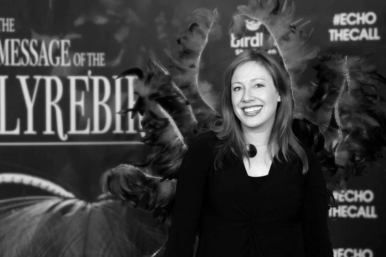 A woman smiling with feathers behind her at an event, with a background that has the text 'THE MESSAGE OF THE' and 'LYREBIRD' visible, along with hashtags #ECHO and #THECALL.