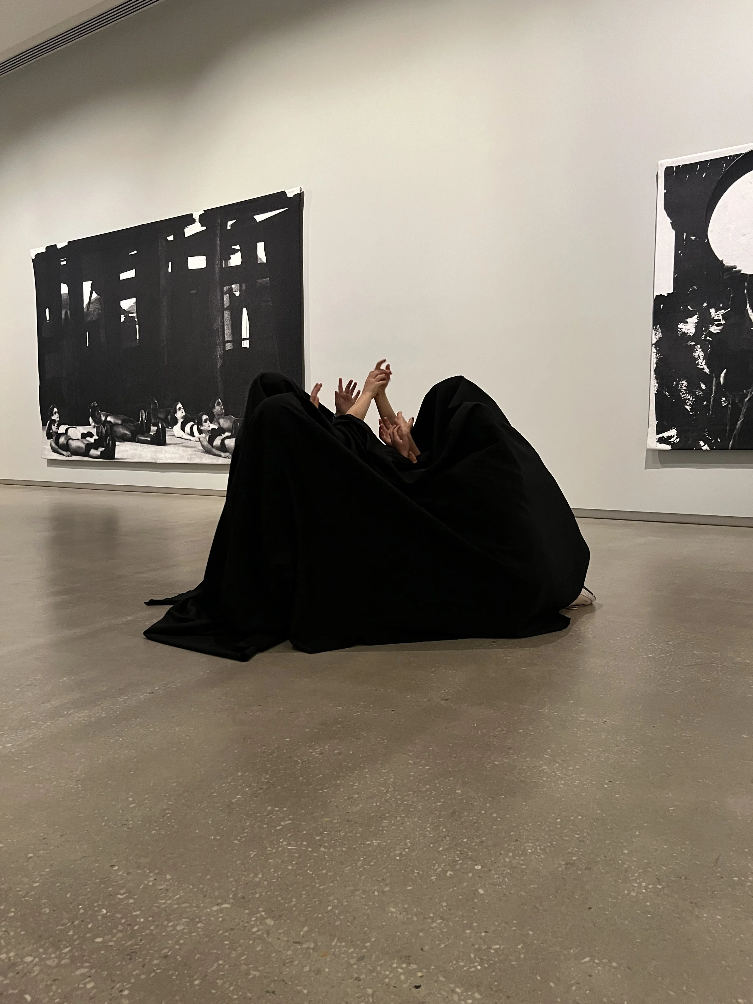 View of a person in black clothing lying on their back on the floor in an art museum, with artwork on the walls behind them.