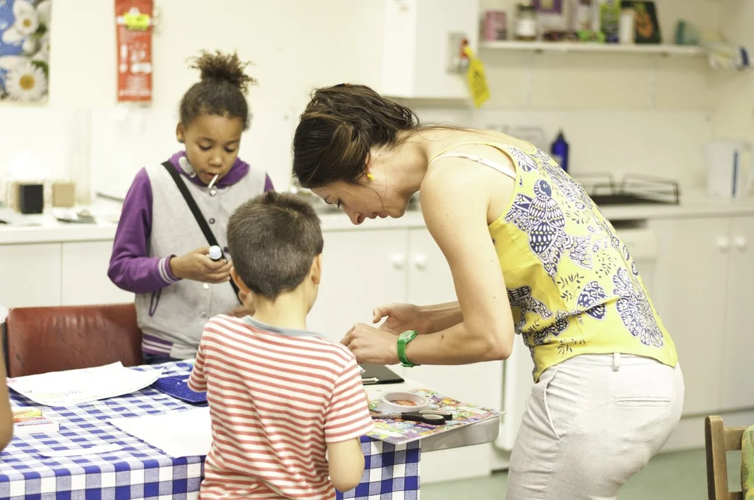 A woman interacts with two children at a table in a room that appears to be a classroom or activity center. One girl holds a glue stick, while a boy looks down at the table. The woman is smiling and leaning toward the boy, engaging with him.