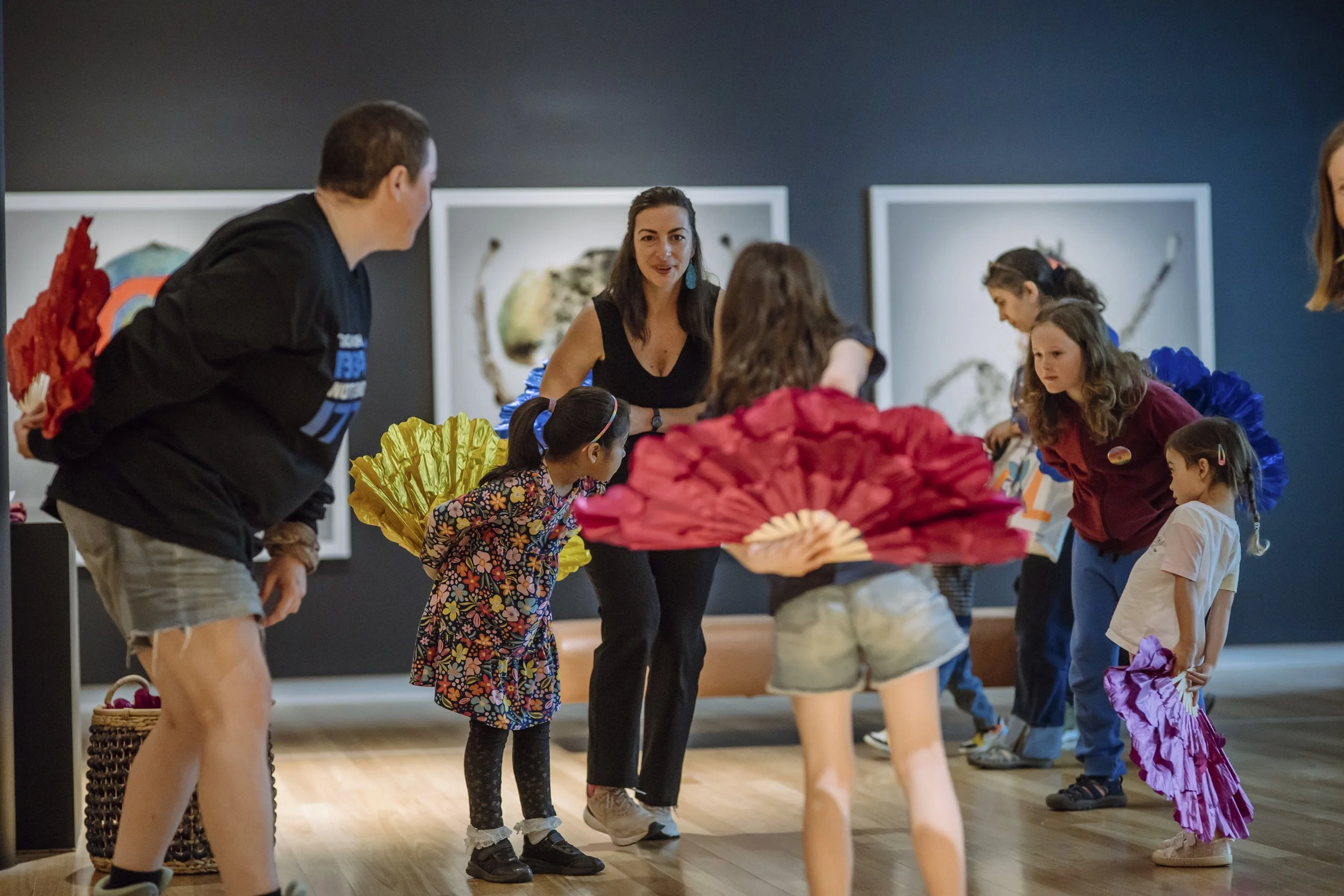 Children and adults participate in a group activity, some holding colorful paper fans, in an indoor space with artwork on the walls.