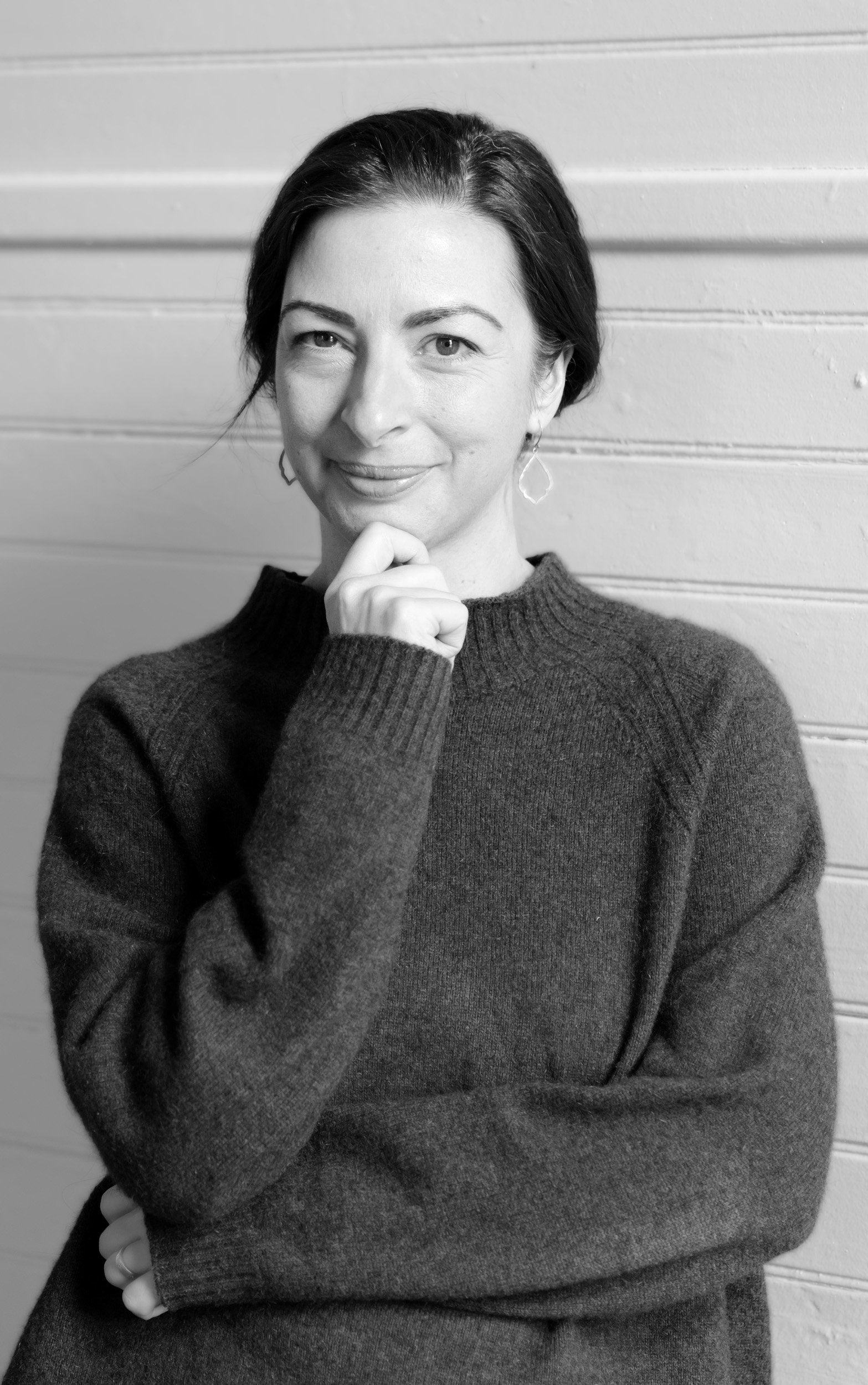 A black and white photo of a woman with short dark hair, wearing a dark long-sleeve sweater, standing against a wooden wall. She is smiling slightly, with one hand near her chin and the other crossed over her stomach.