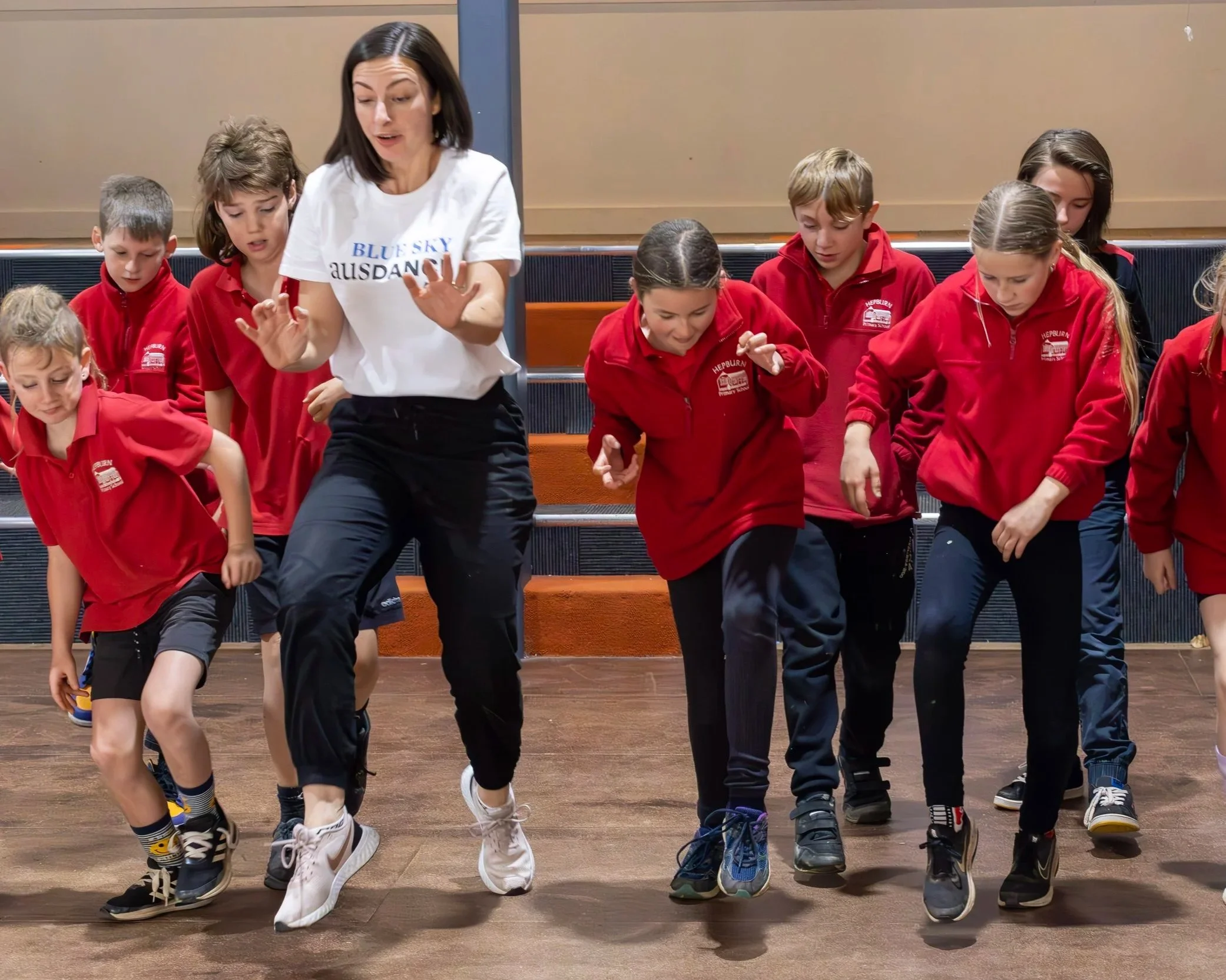 Group of children in red school uniforms standing on benches, engaged in a dance or activity, led by a woman with shoulder length dark hair wearing a white T-shirt, in an indoor setting.