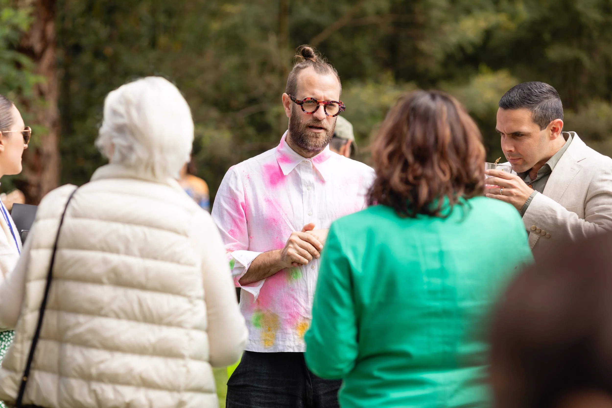 A man with a top knot and colorful glasses speaking to a group of people outdoors, some wearing jackets and one with gray hair, in a wooded area.