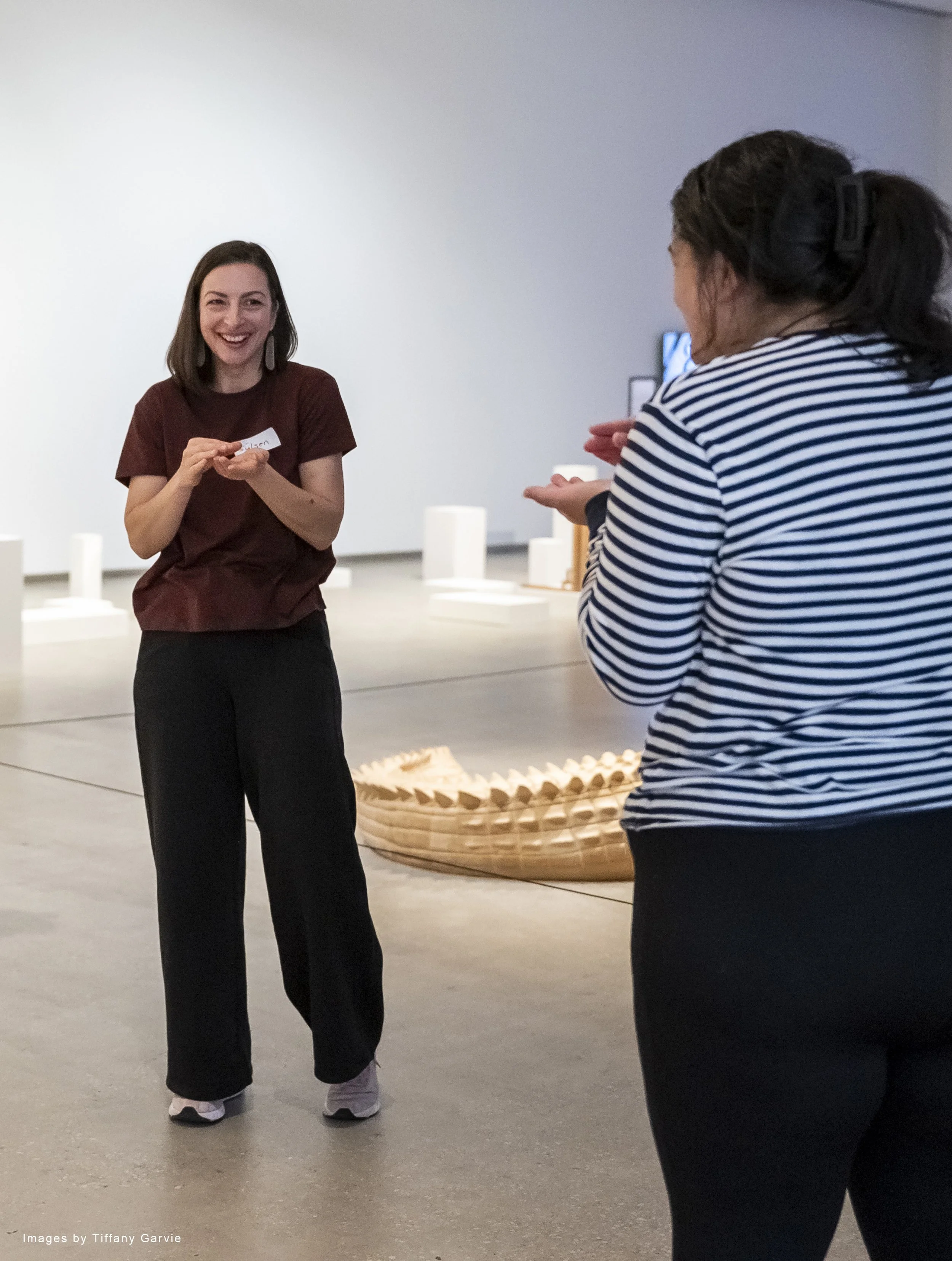 Two women engaging in conversation in an art gallery. One woman is facing the camera with a smile, wearing a maroon top and black pants. The other woman, with her back to the camera, is wearing a striped shirt and has her hair in a ponytail with a black hair clip. There is a large basket and aTV or monitor in the background.
