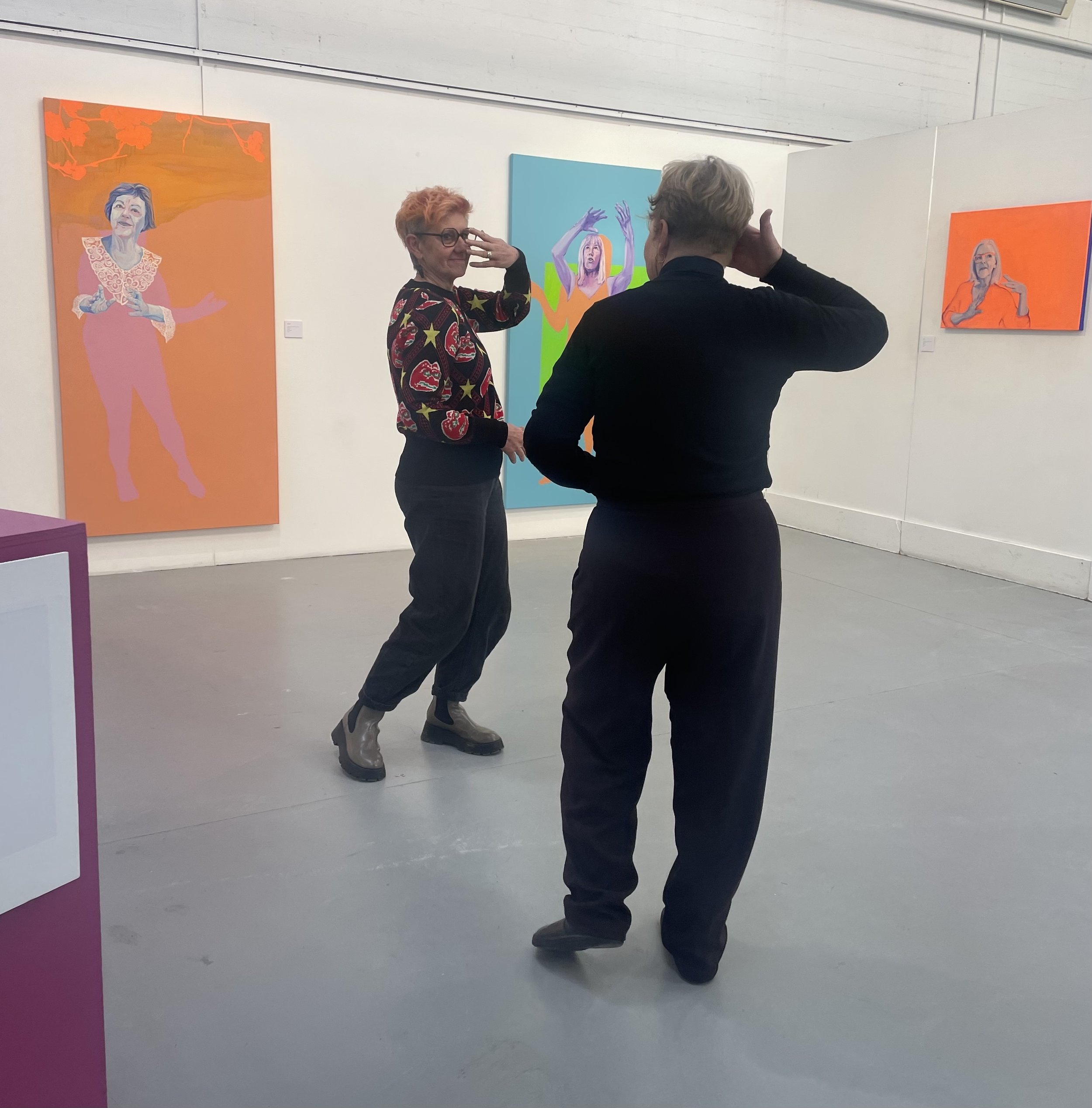 Two women are conversing in an art gallery, surrounded by colorful artwork on the walls.