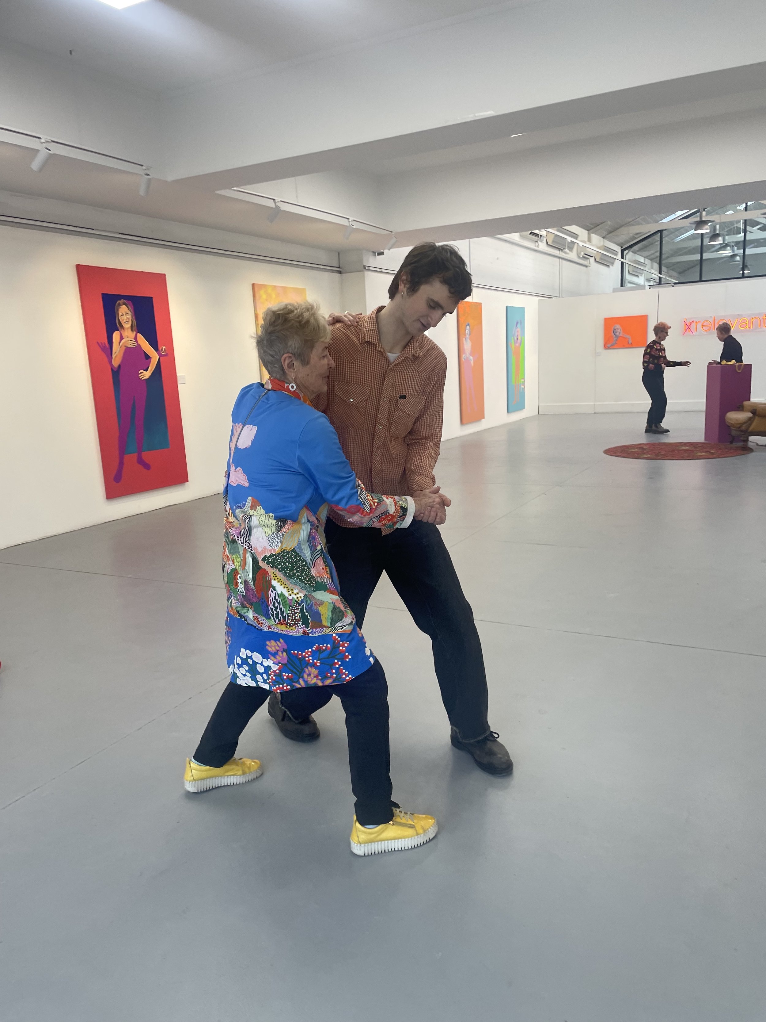 Two people are dancing together in an art gallery. One is wearing a colorful patterned jacket and yellow sneakers, while the other is dressed in a brown checkered shirt and black shoes. They appear to be engaged in a dance move or playful interaction
