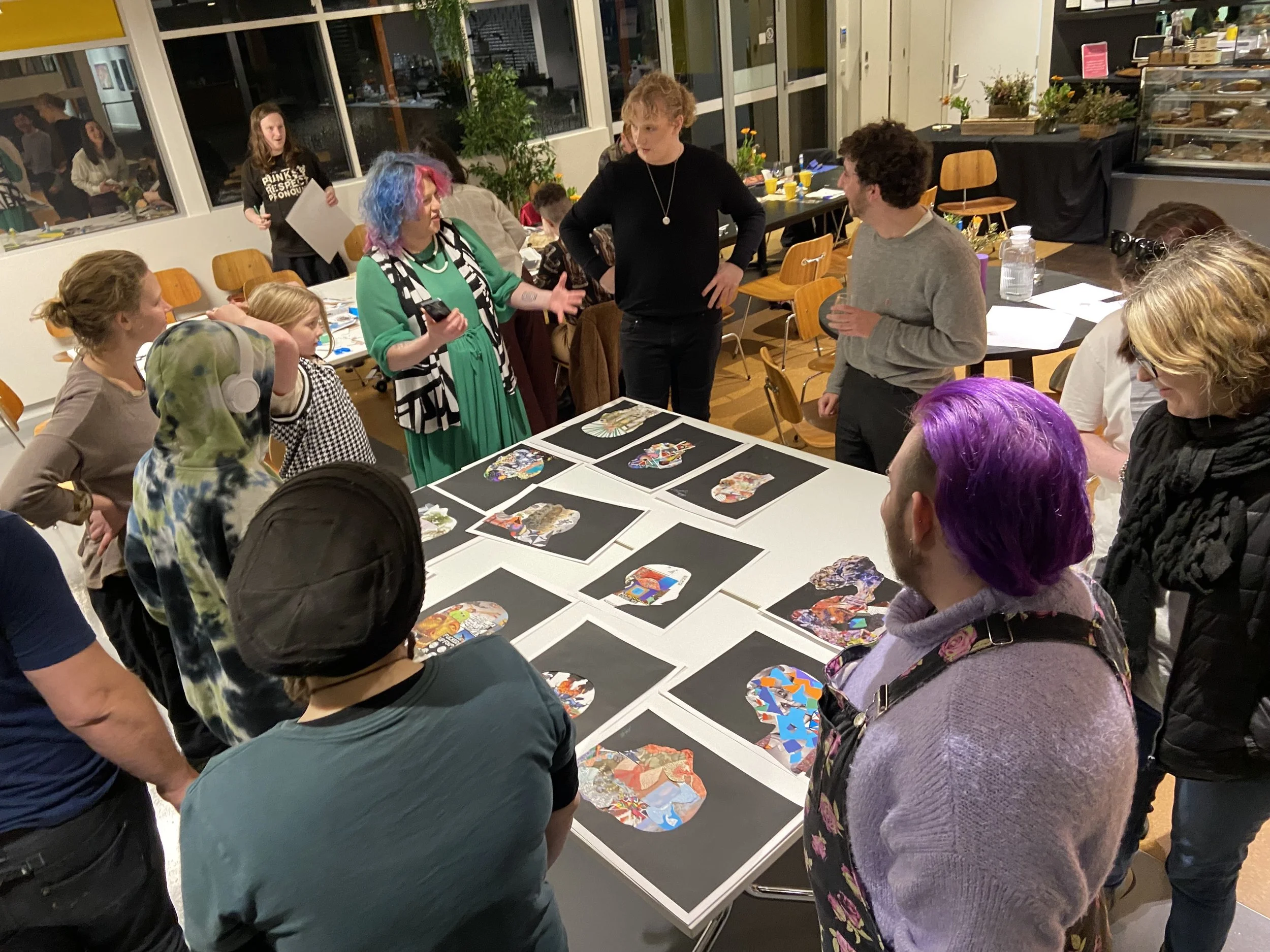 A group of people gathered around a table with colorful artwork or collage pieces laid out for display or discussion. Some people are engaged in conversation, while others are observing the artwork. In the background, there are windows, and a few individuals can be seen through them, along with plants and a buffet area.