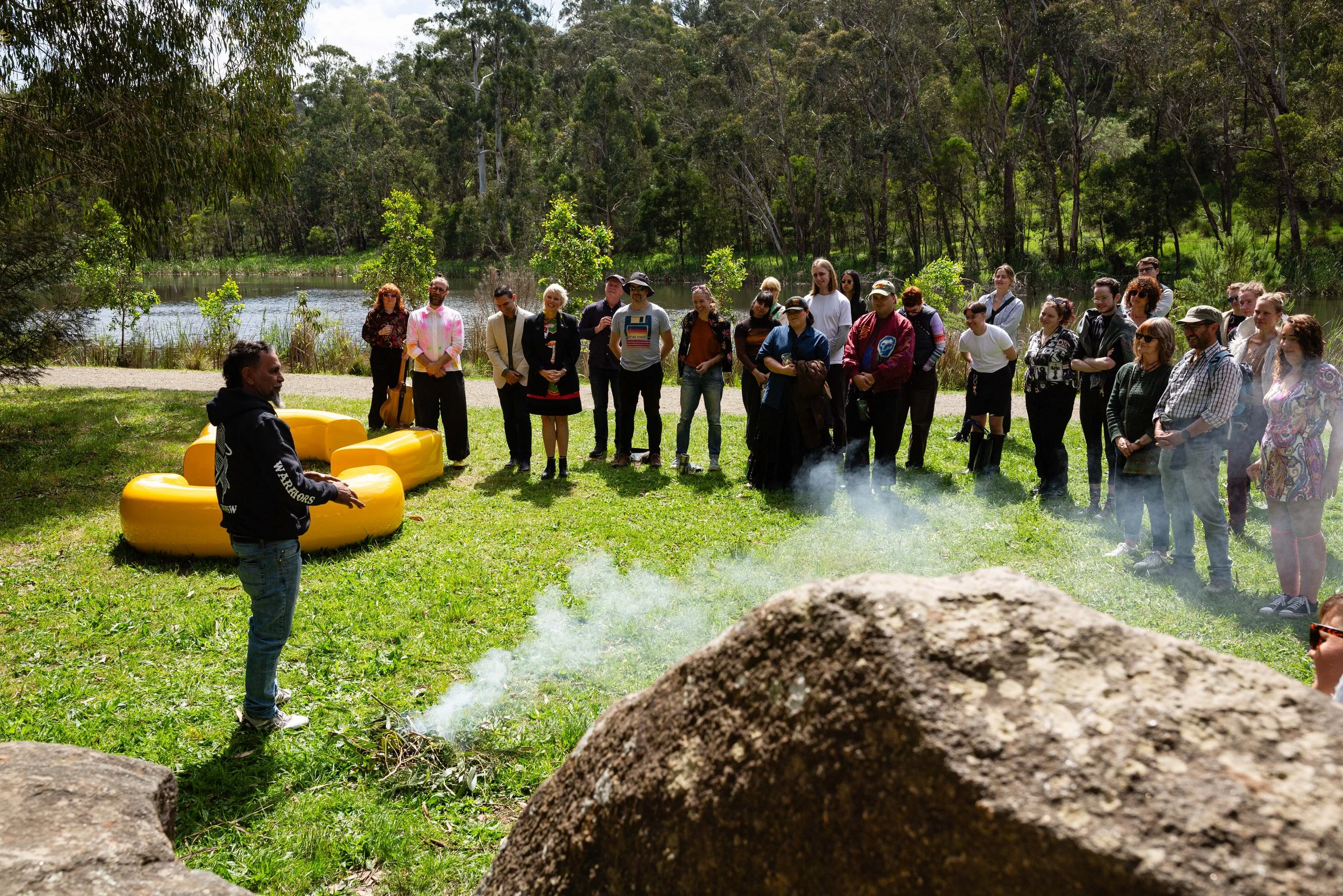 Group of people gathered outdoors near a pond, listening to a person speaking in front of them. The setting is lush with green trees and grass, and some smoke or steam is visible on the ground.