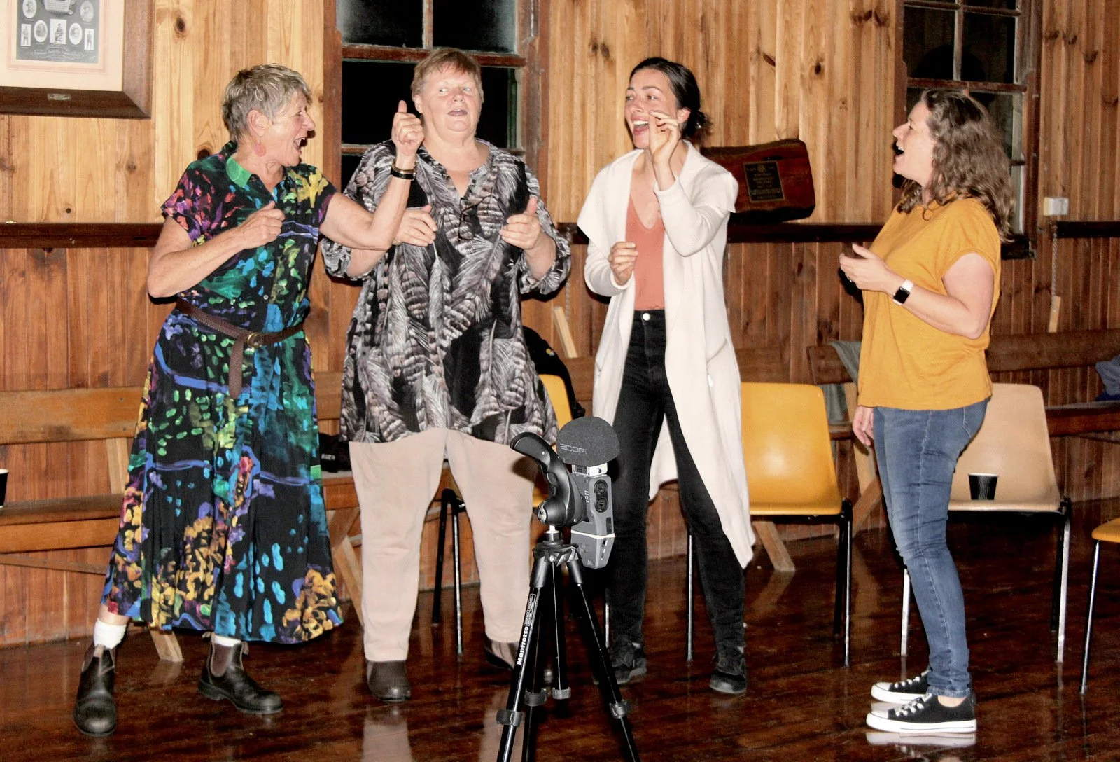 Four women standing together in a wooden-paneled room, engaging in a lively conversation or celebration, with a camera on a tripod in front of them.