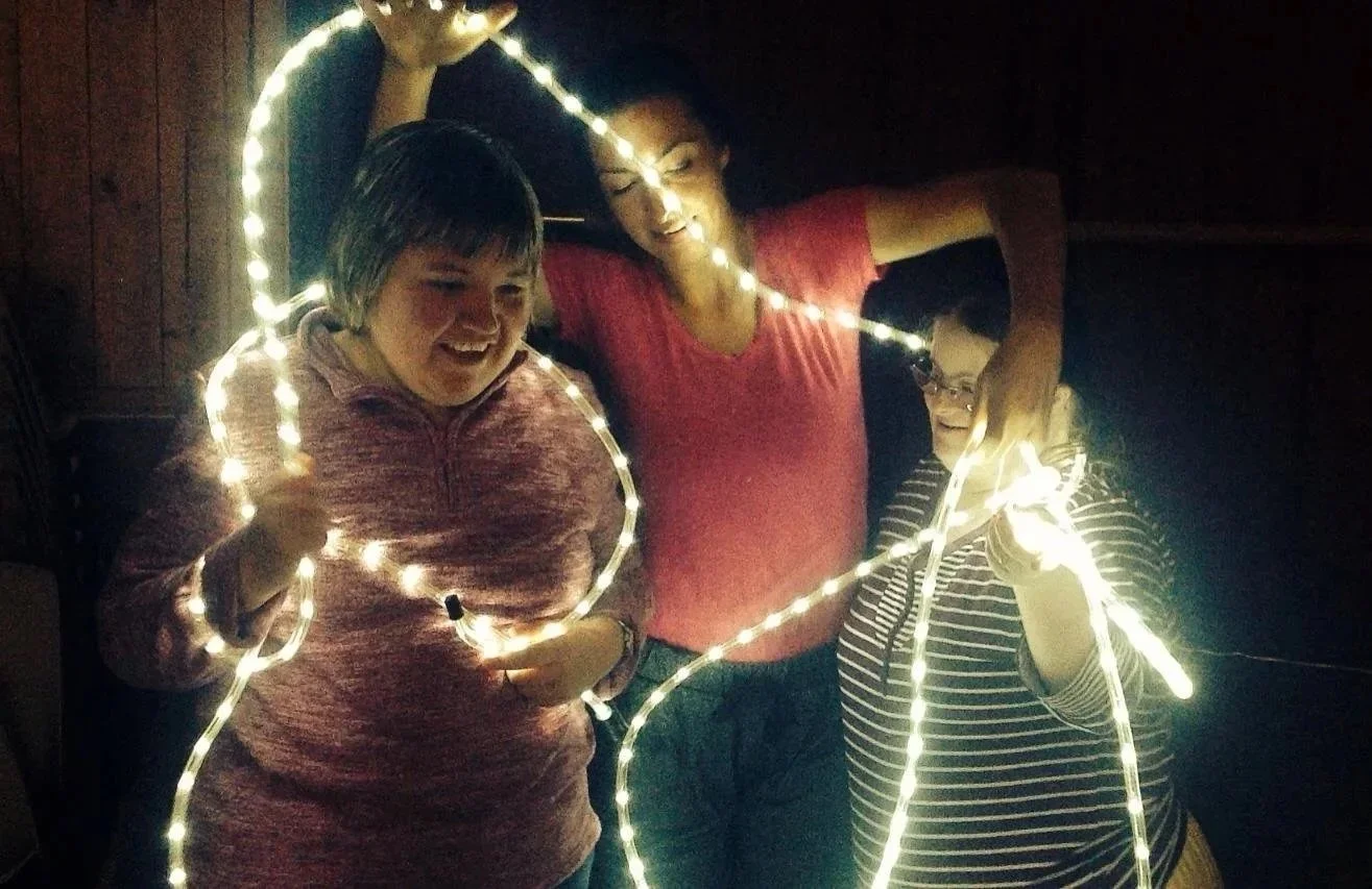 Three people are smiling and holding a string of glowing lights, creating a festive atmosphere in a dark room.