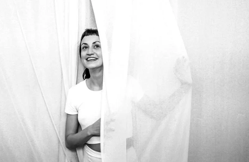 A young woman with dark hair and a white t-shirt peeking out from behind a curtain, smiling.