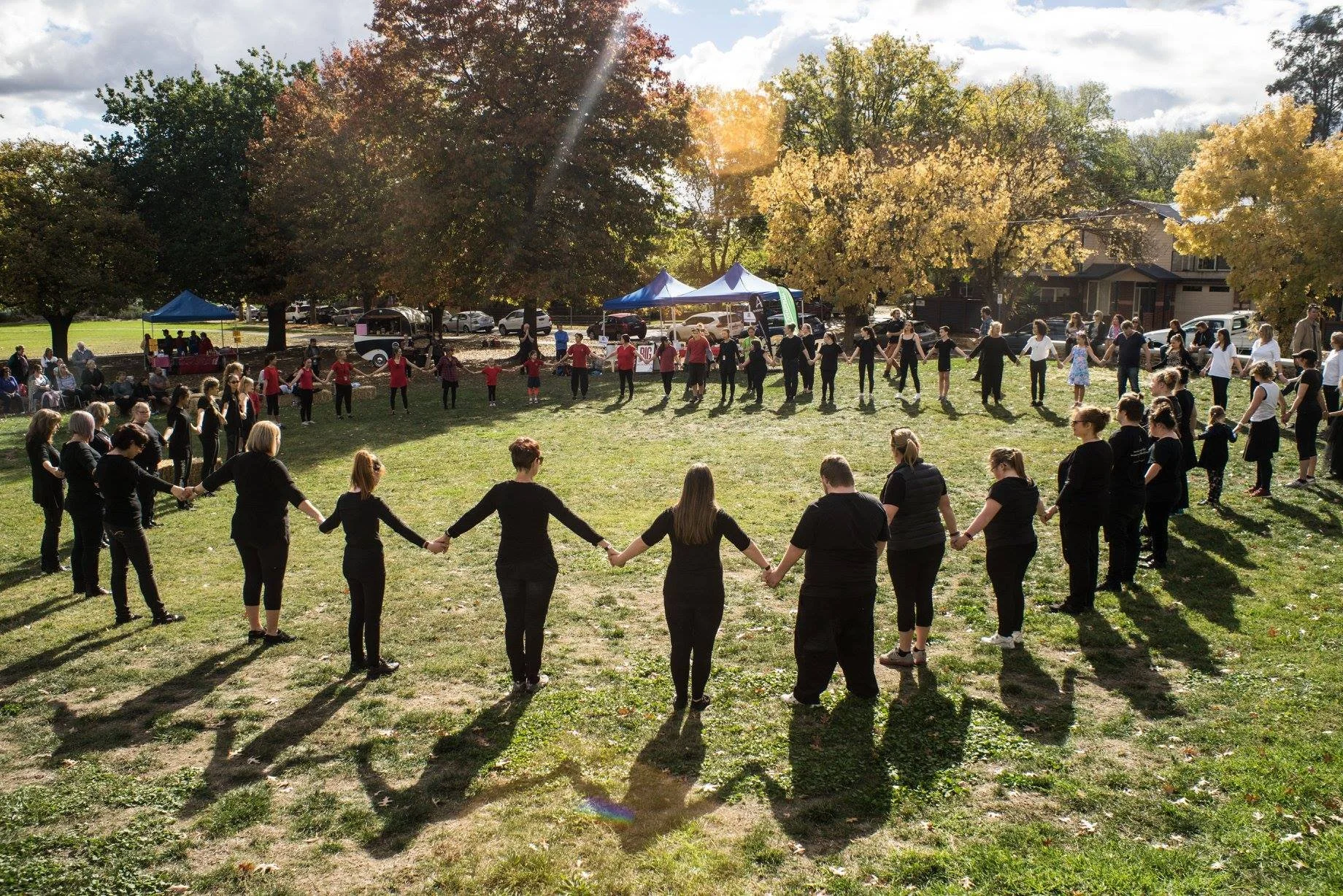 A large group of people holding hands in a circle in a park, with trees and tents in the background during a sunny day.