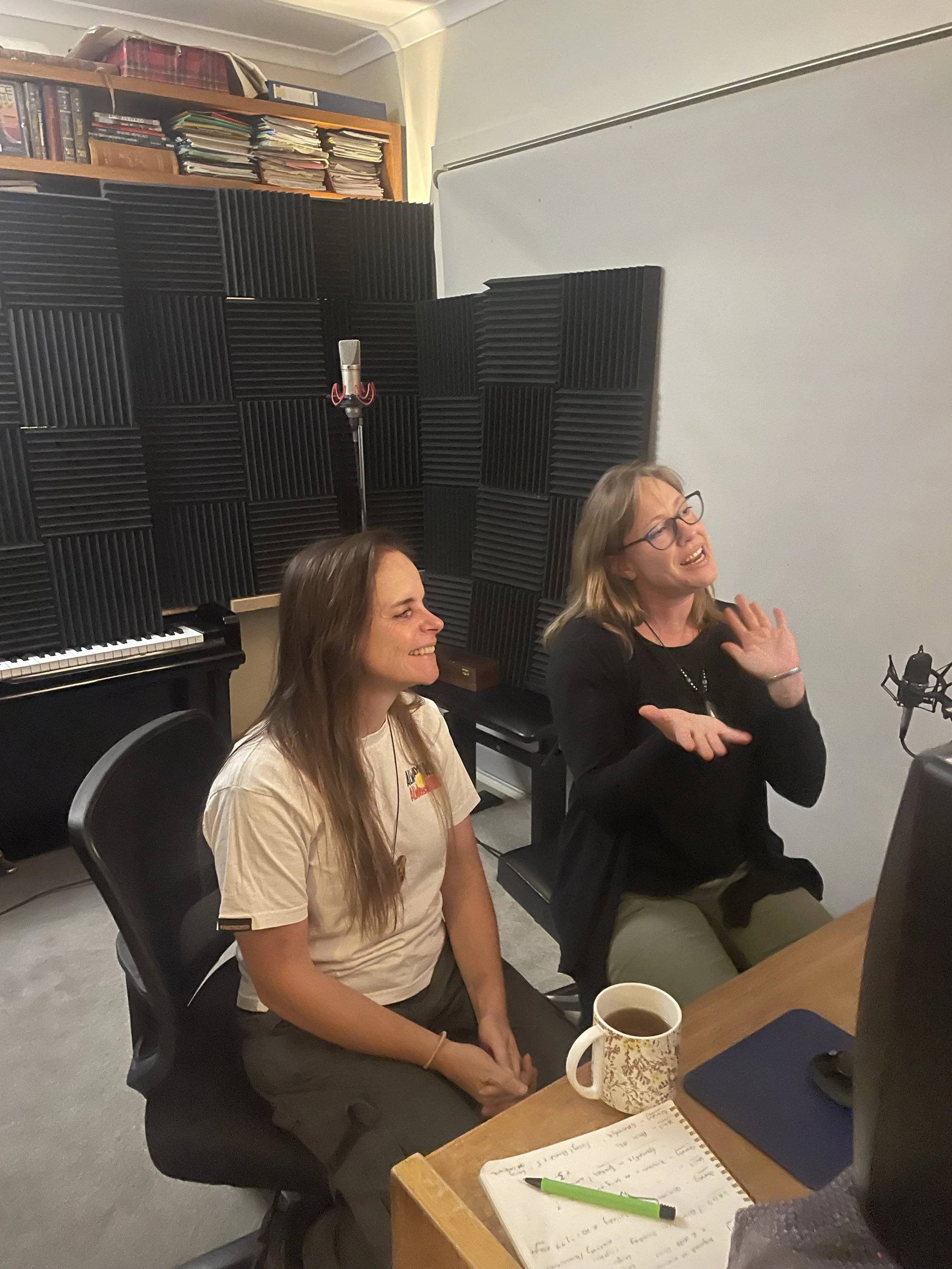 Two women sitting at a table in a recording studio, laughing and talking. One woman is wearing glasses and a black shirt, the other has long hair and is wearing a beige shirt. There are headphones, a mug, a notebook, and a pen on the table, with a keyboard and soundproofing foam in the background.
