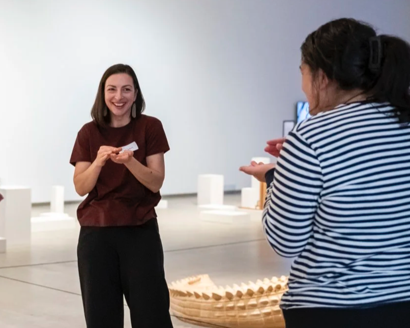 Two women having a conversation in an art gallery, with one woman smiling and holding her hands together, and the other, with curly hair tied back, gesturing with her hands, in front of white display pedestals and artwork.