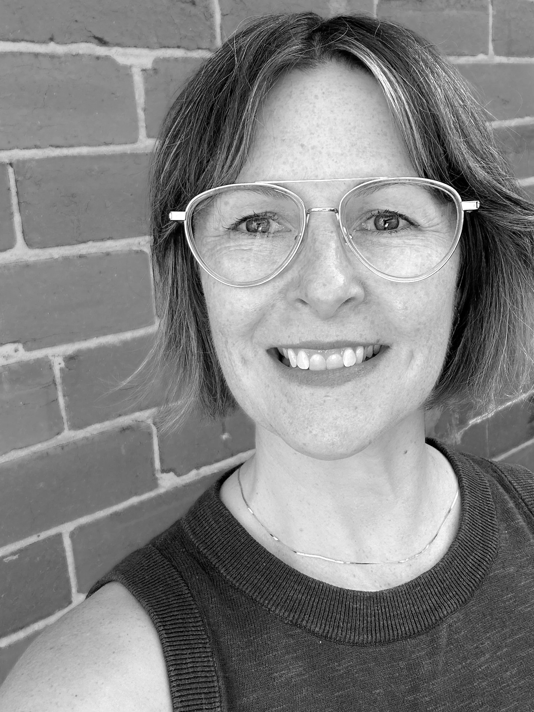 Black and white photo of a smiling woman with short hair wearing glasses, standing against a brick wall.