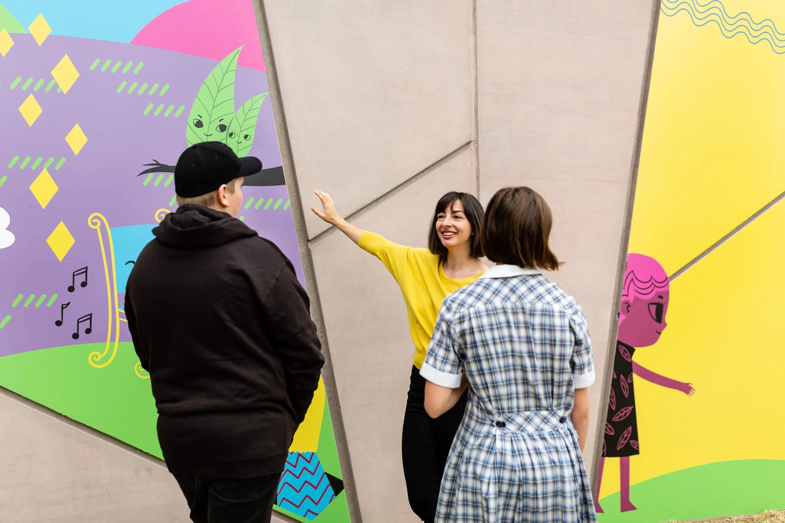 A woman in a yellow shirt talking to two people, with colorful murals in the background depicting cartoonish characters and patterns.