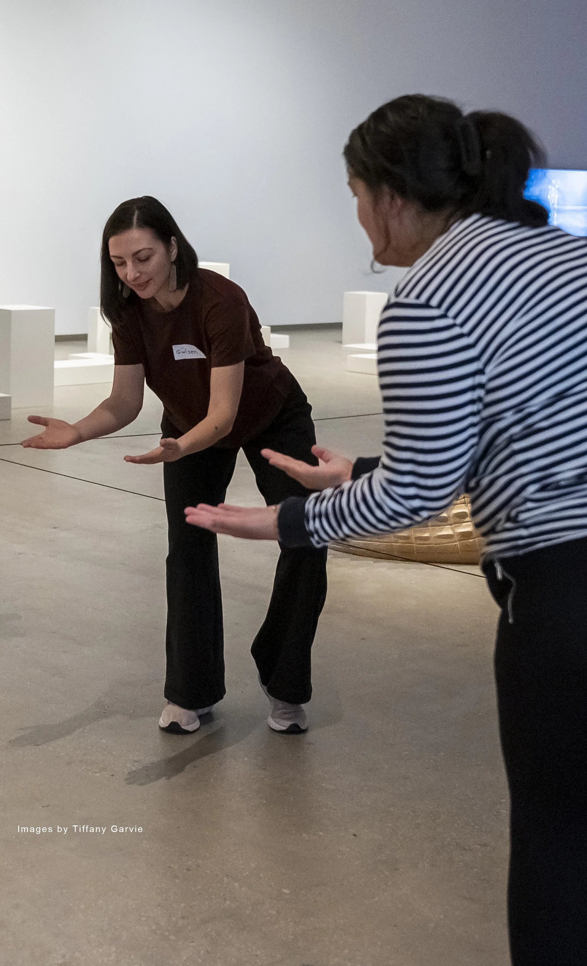 Two women performing a dance or movement exercise in an indoor space with white walls and minimal decor. One woman has dark hair, is wearing a striped shirt, and the other has dark hair, is wearing a dark-colored top with a name tag, and black pants.