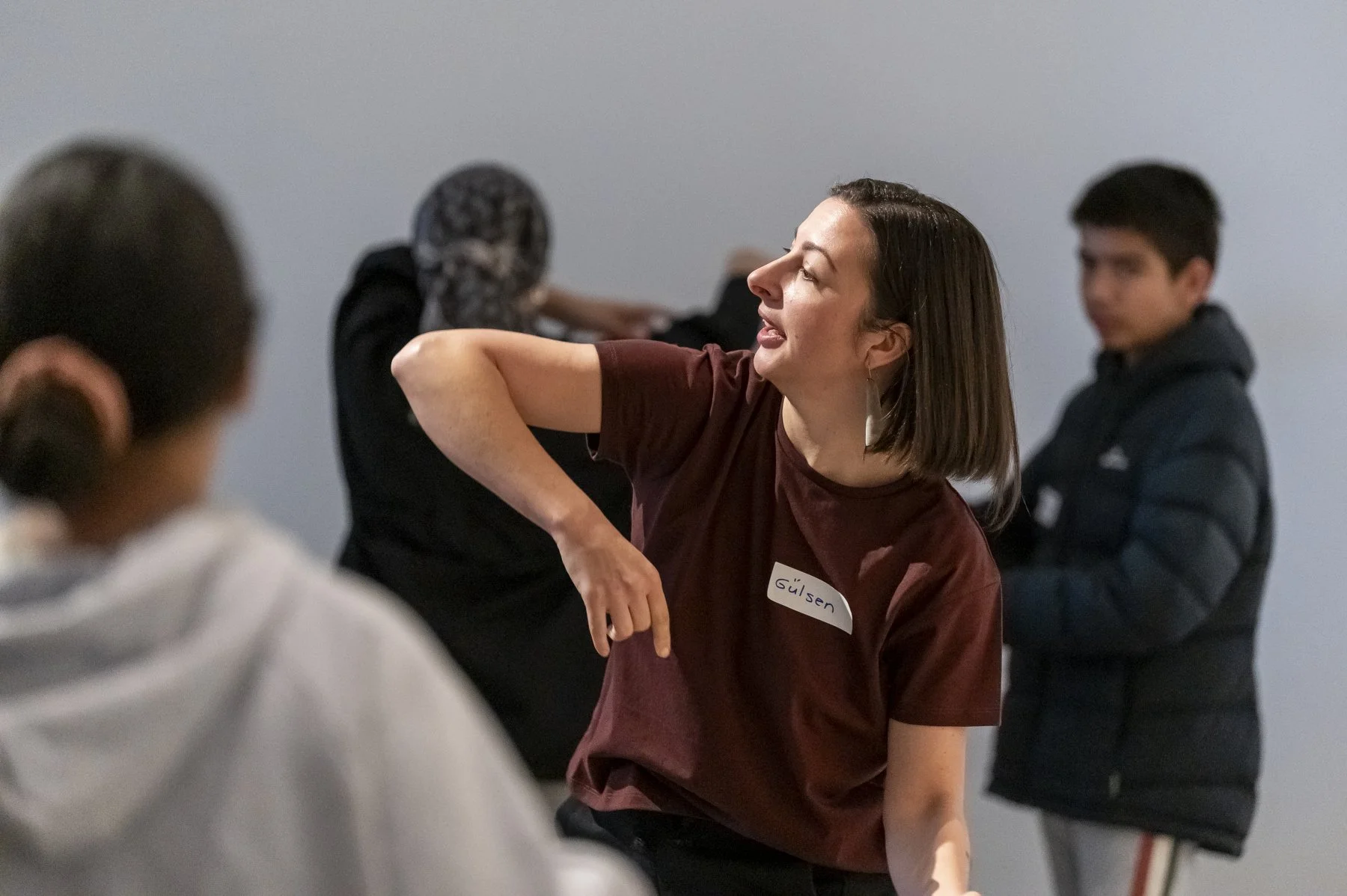 A woman with shoulder-length brown hair, wearing a maroon shirt and a name tag that reads 'Gülsen,' is gesturing animatedly with her right hand while speaking to a group of people in an indoor setting. Several other people are visible, including a woman with braided hair and hijab, a young man in a striped jacket, and a person with glasses and a hoodie, all engaged in conversation.