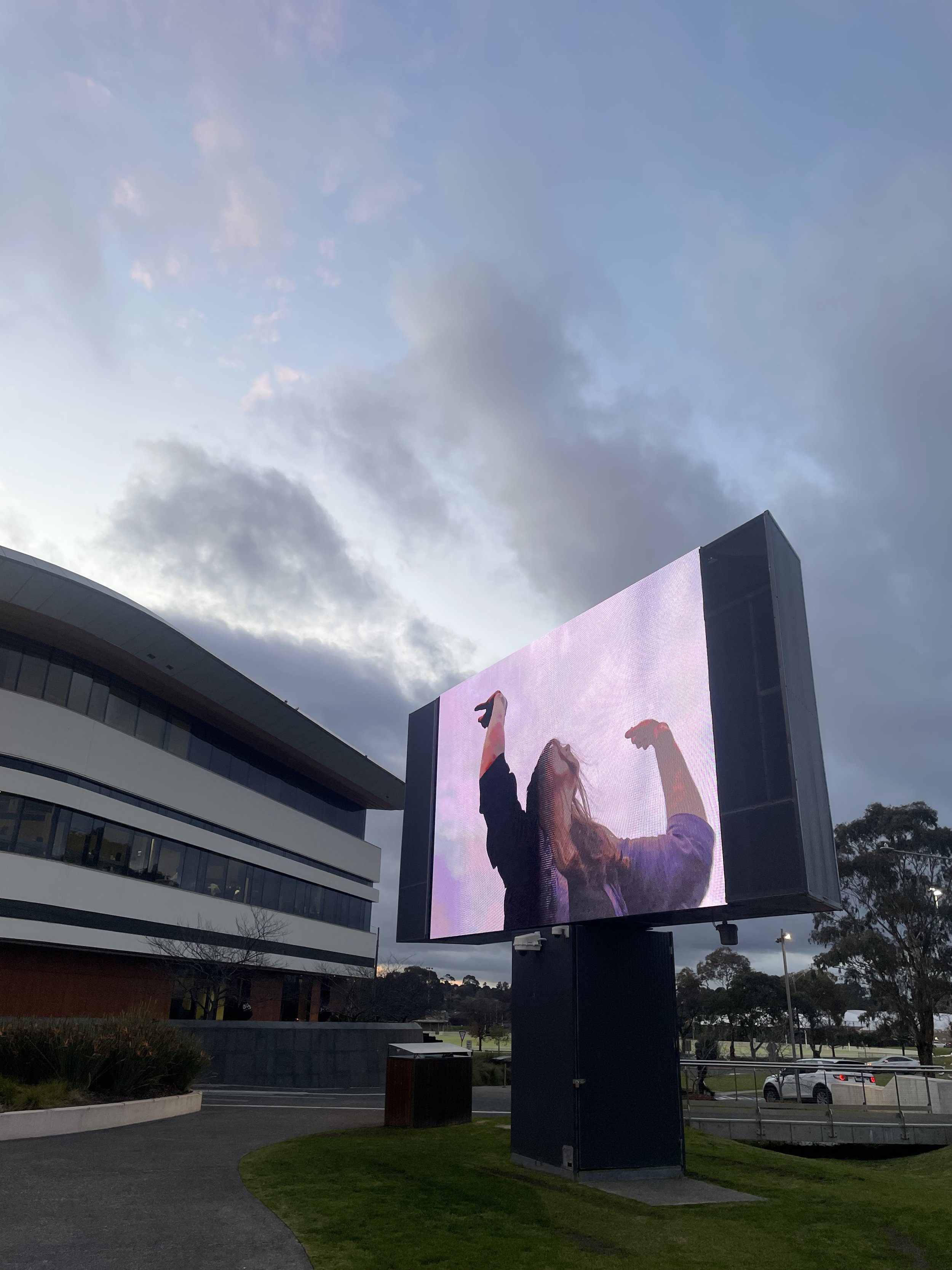 A large outdoor digital billboard showing a woman with long hair posing with arms raised and fists clenched in a celebratory manner. The billboard is situated near a curved building with multiple floors, and the sky is partly cloudy during the evening or late afternoon.