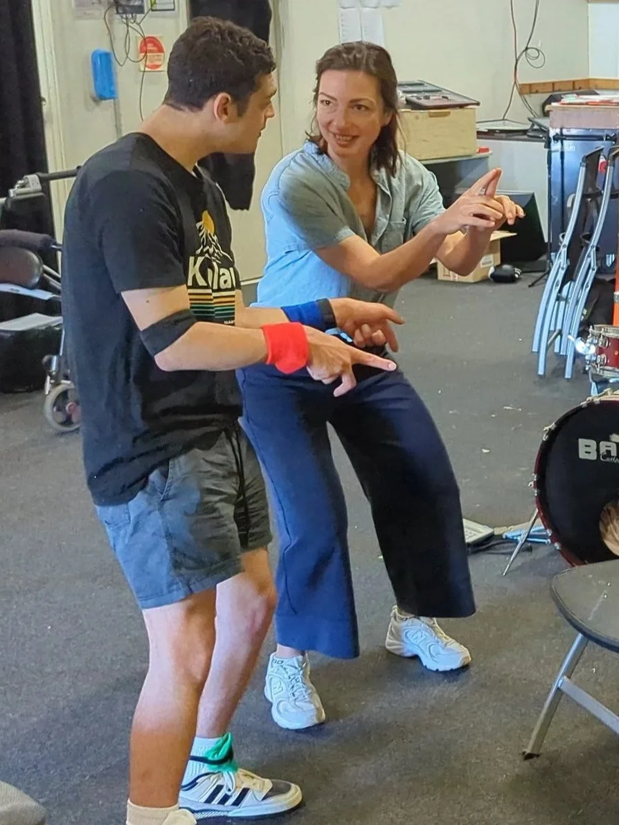 Two people, a man and a woman, are interacting and pointing at something together in a casual indoor setting, possibly a rehearsal or practice space.