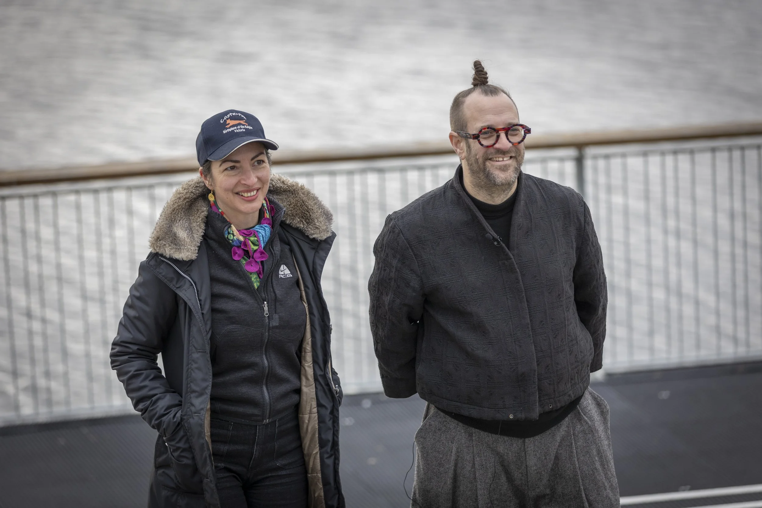 A woman and a man walking outdoors near a body of water. The woman is wearing a dark jacket with a fur-lined collar, a colorful scarf, and a navy cap. The man is wearing glasses with a tortoiseshell pattern, a dark textured jacket, and gray pants. They are smiling and looking ahead.