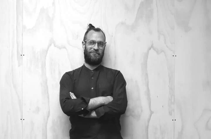 Black and white photo of a man with glasses and a beard, standing with arms crossed against a wood-paneled wall. He is wearing a button-up shirt.