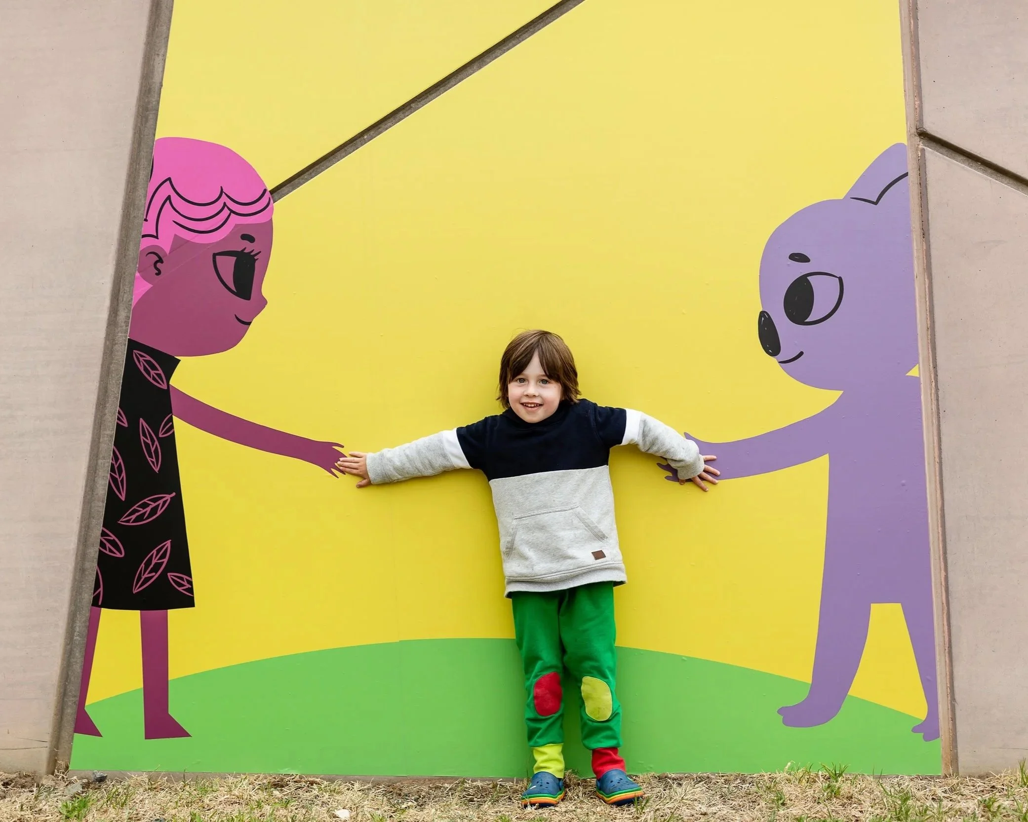 A young boy with brown hair standing in front of a colorful mural with cartoon faces, arms outstretched. The mural features a pink character on the left and a purple character on the right, both smiling.