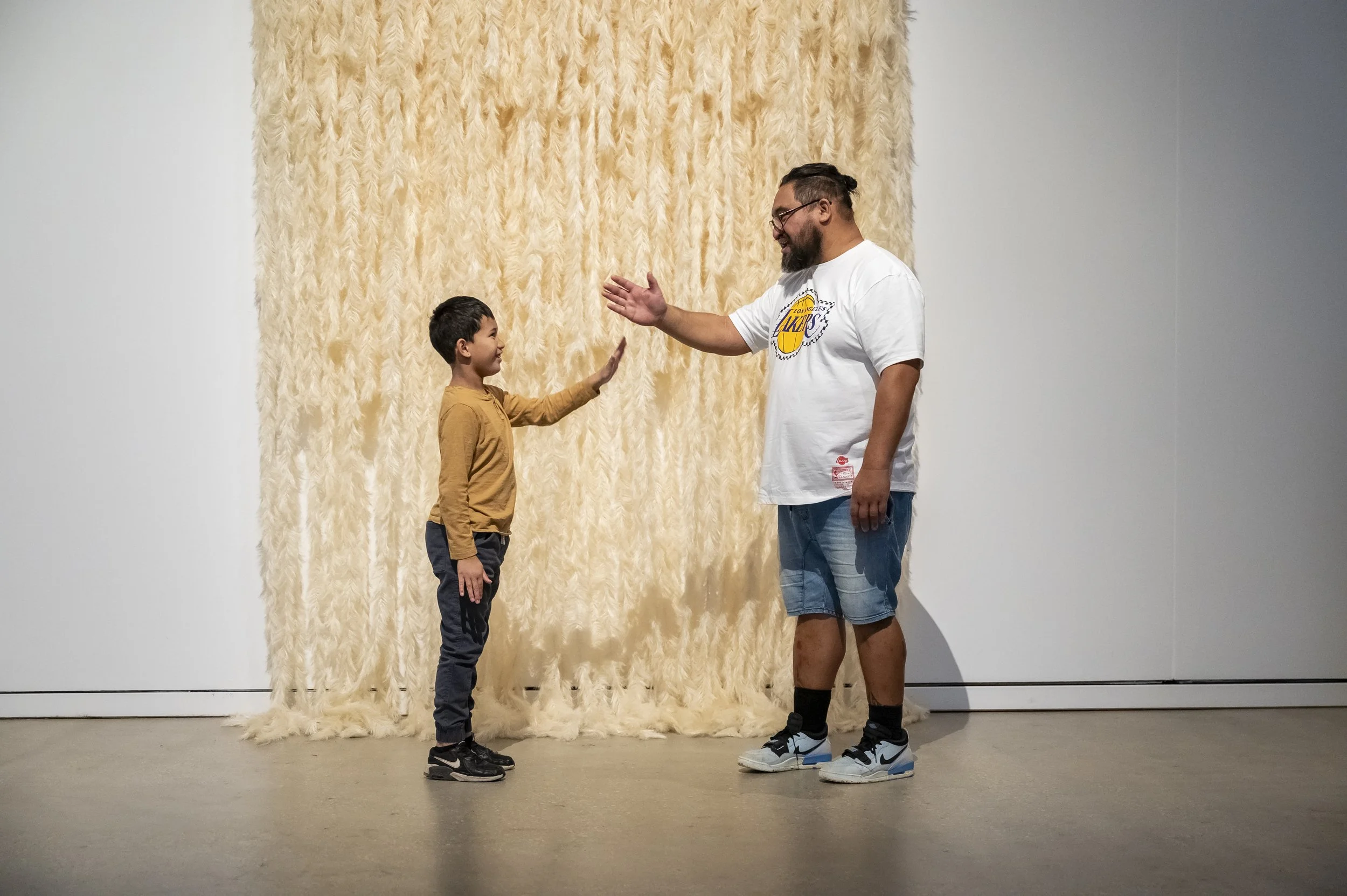 A man and a boy standing in front of a large cream-colored, fluffy textured backdrop, high-fiving each other. The man is wearing a white T-shirt with a Lakers logo, shorts, and sneakers, while the boy is dressed in a brown shirt, pants, and sneakers.
