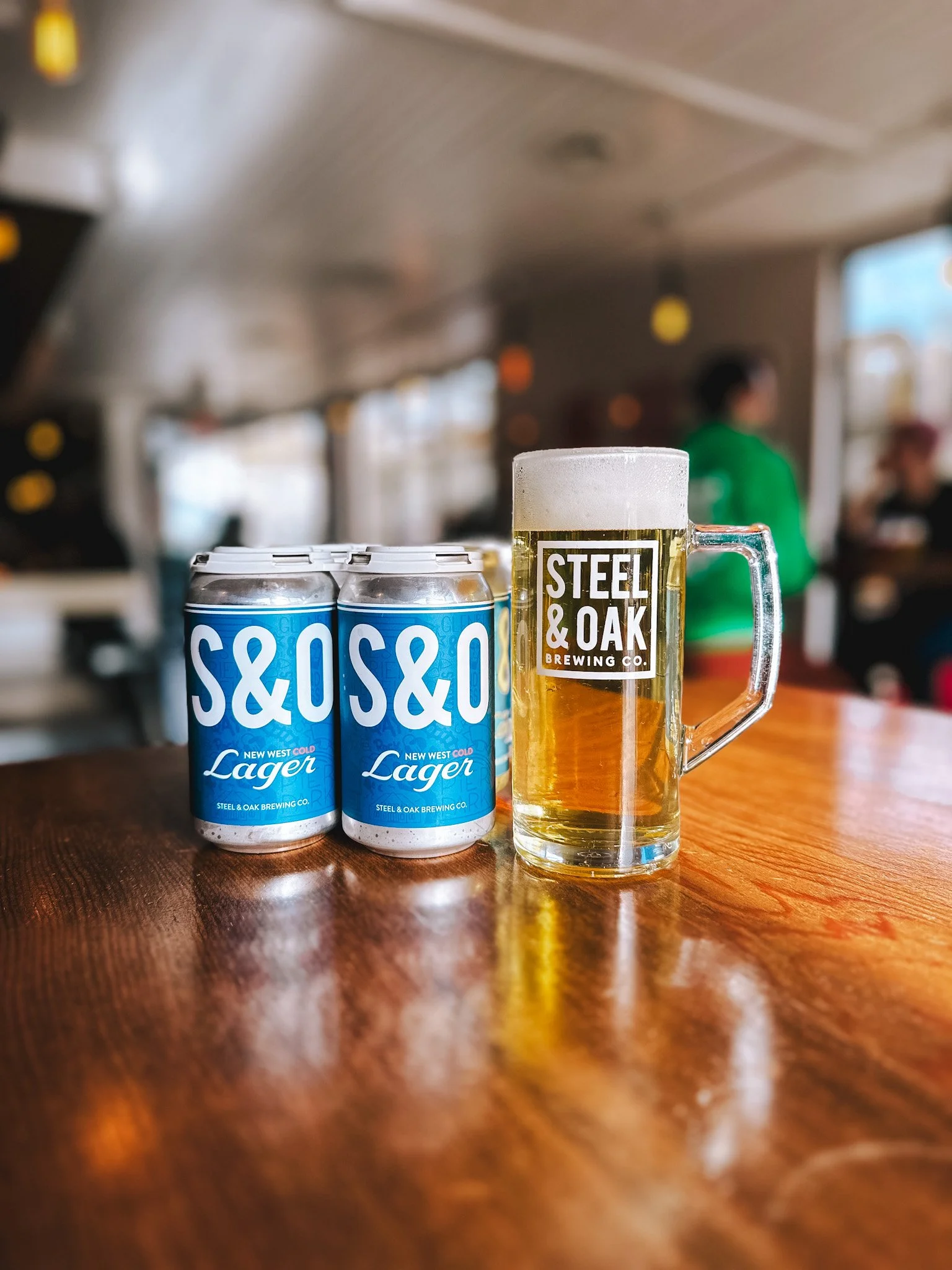 Three cans of Steel & Oak lager beer beside a glass of beer on a wooden table